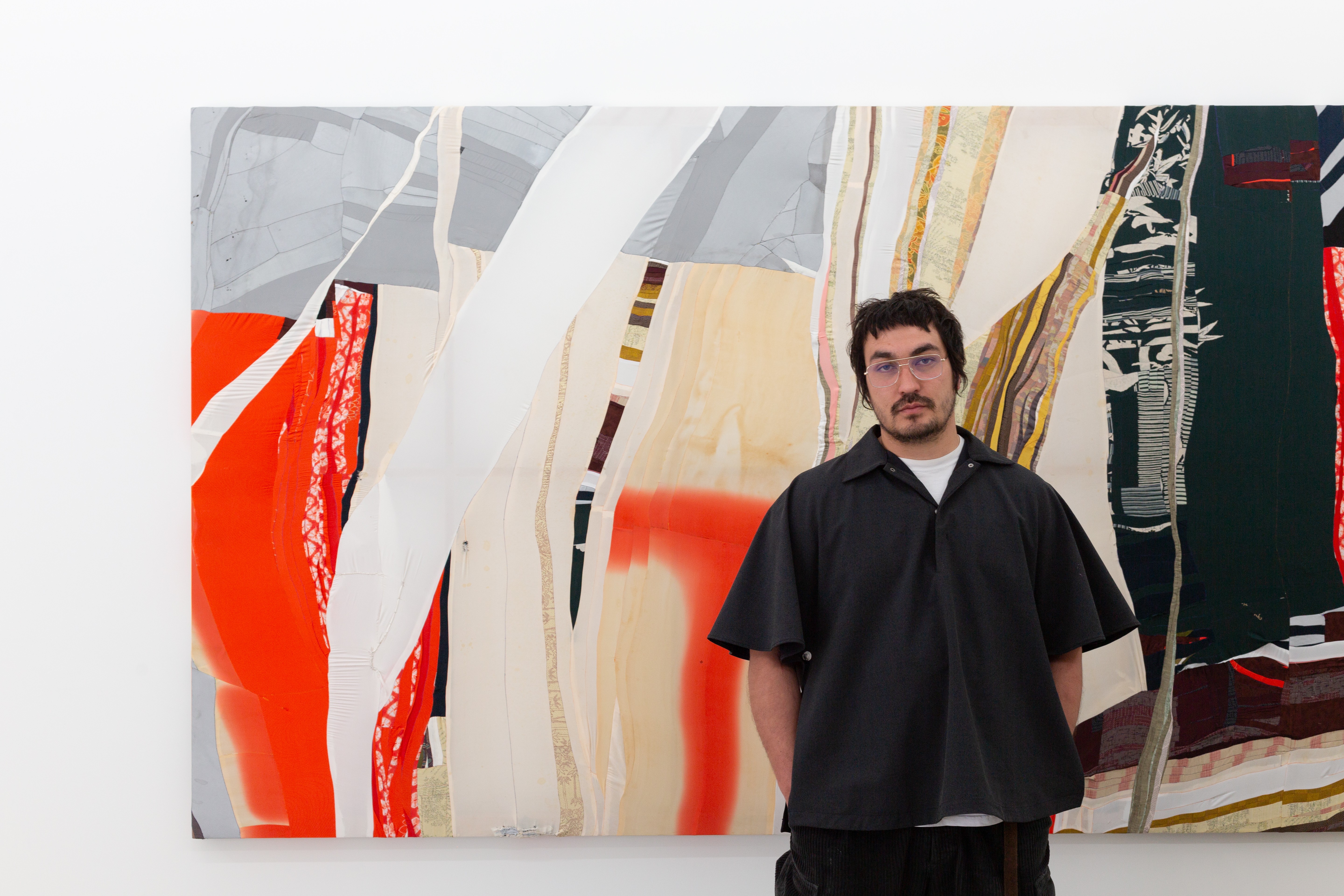 Dark haired man with glasses stands solemnly in front of a bright and large abstract artwork