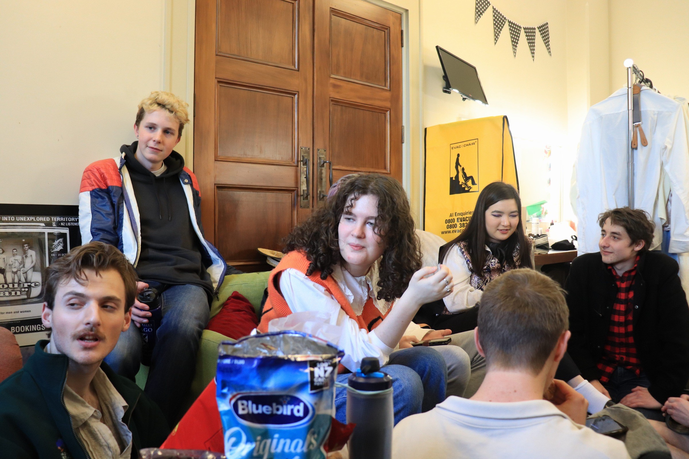 A group of people sitting on a couch and on the floor around it. A packet of crisps is visible.