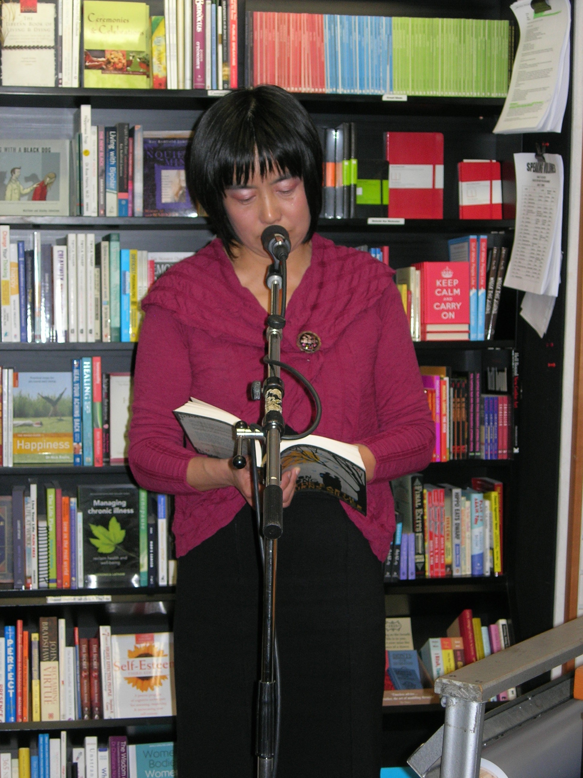A woman wearing a burgundy knit reading from her book in front of a microphone in a bookshop