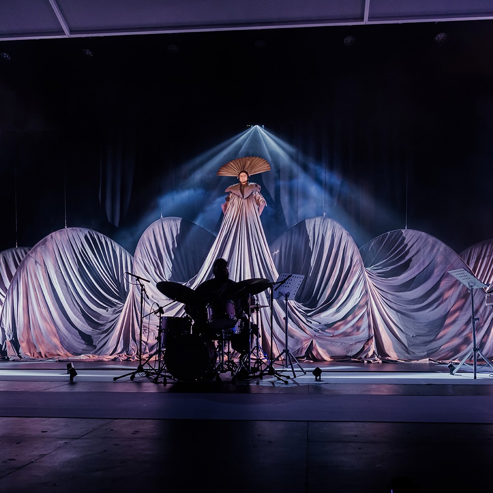 Pink and purple lighting on a stage illuminates a billowing oversized costume worn by a performer