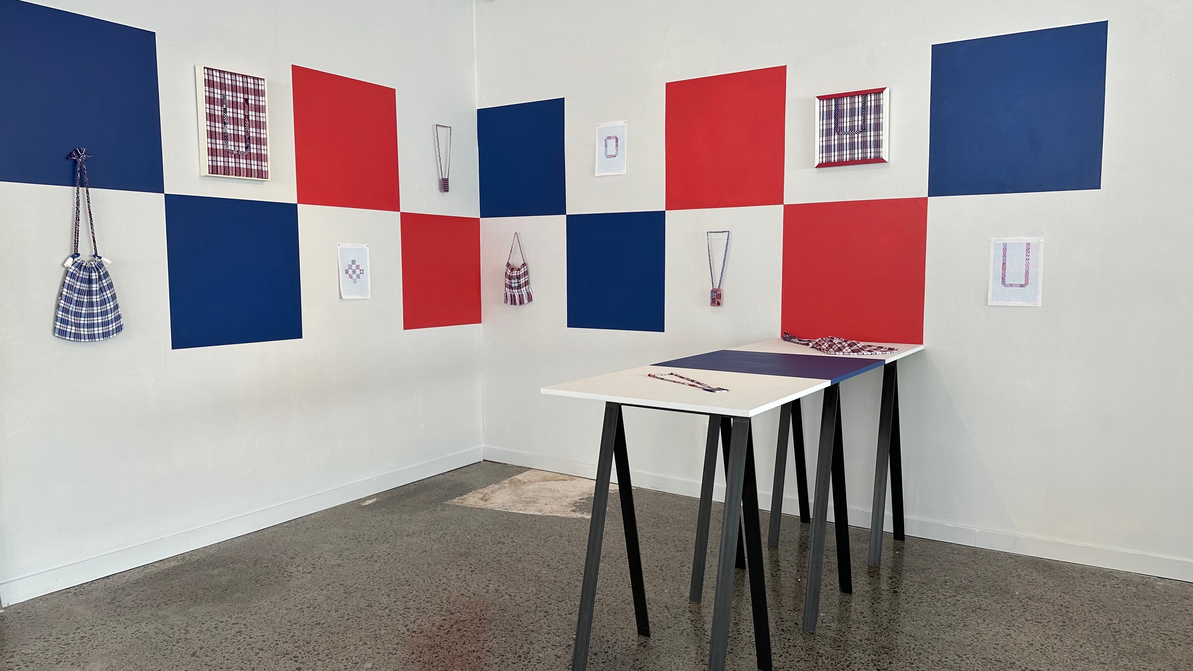 Small embroidered plaid works and matching woven bags are hung on red, white and blue squares painted on a white wall. A small table has two additional works lying on it.