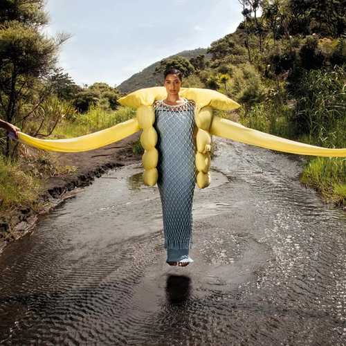 A South Asian woman stands in a river, wearing a blue and net dress with a yellow puffer jacket with extra long sleeves.