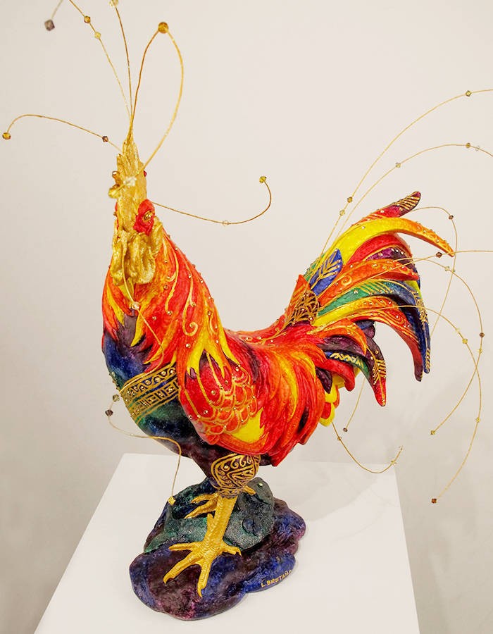 A sculpture of a colourful rooster with various gold metal wiring attached to its body - the gold wiring has colourful crystals attached to them