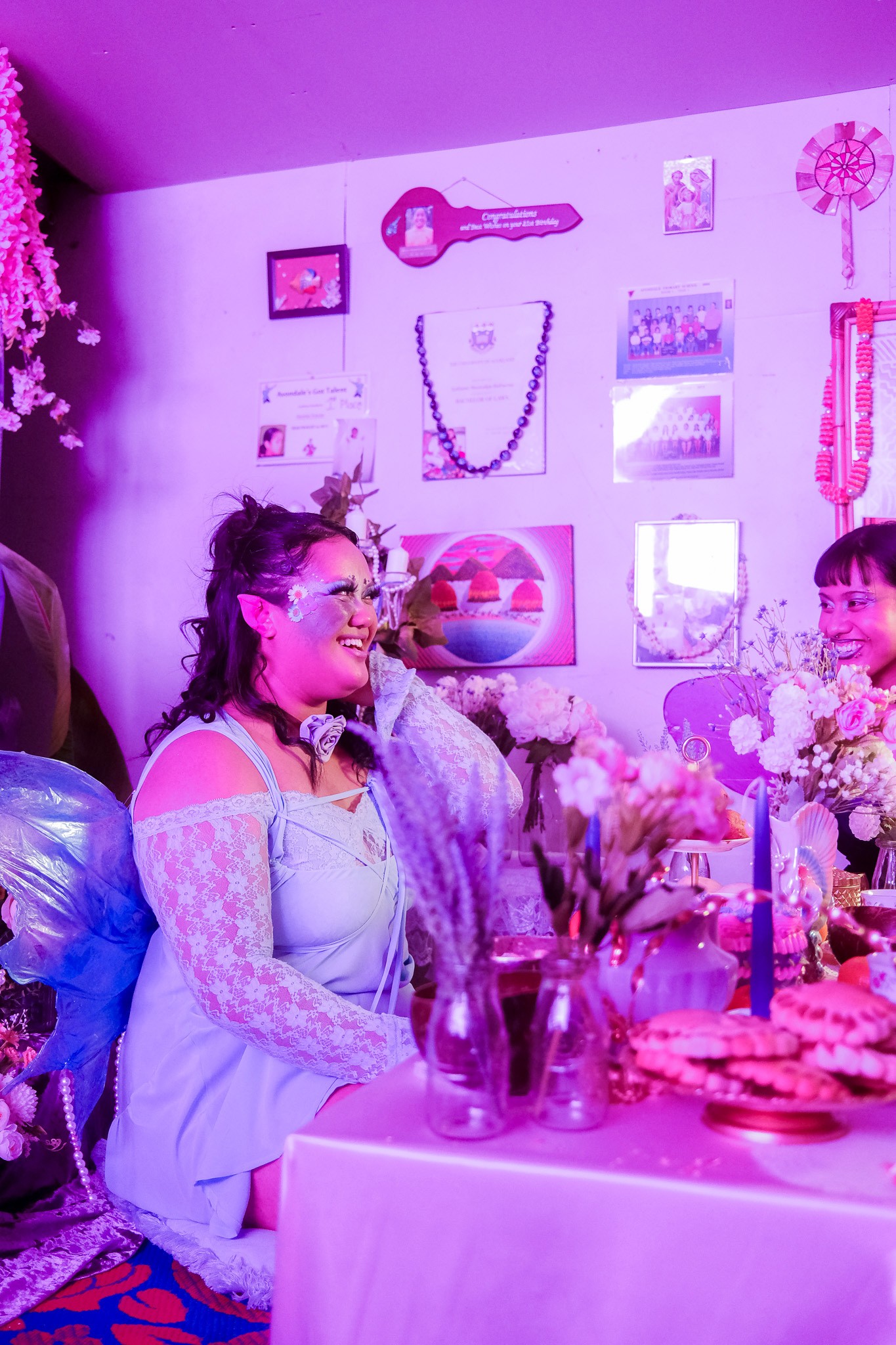 A smiling person in a fashionable powder blue dress wearing blue faerie wings, elf ears and daisies is seated in a dining table that is heaving with various props like candles, glass jars and cookies. The lighting is a soft lavender and gives the photo an ethereal appeal