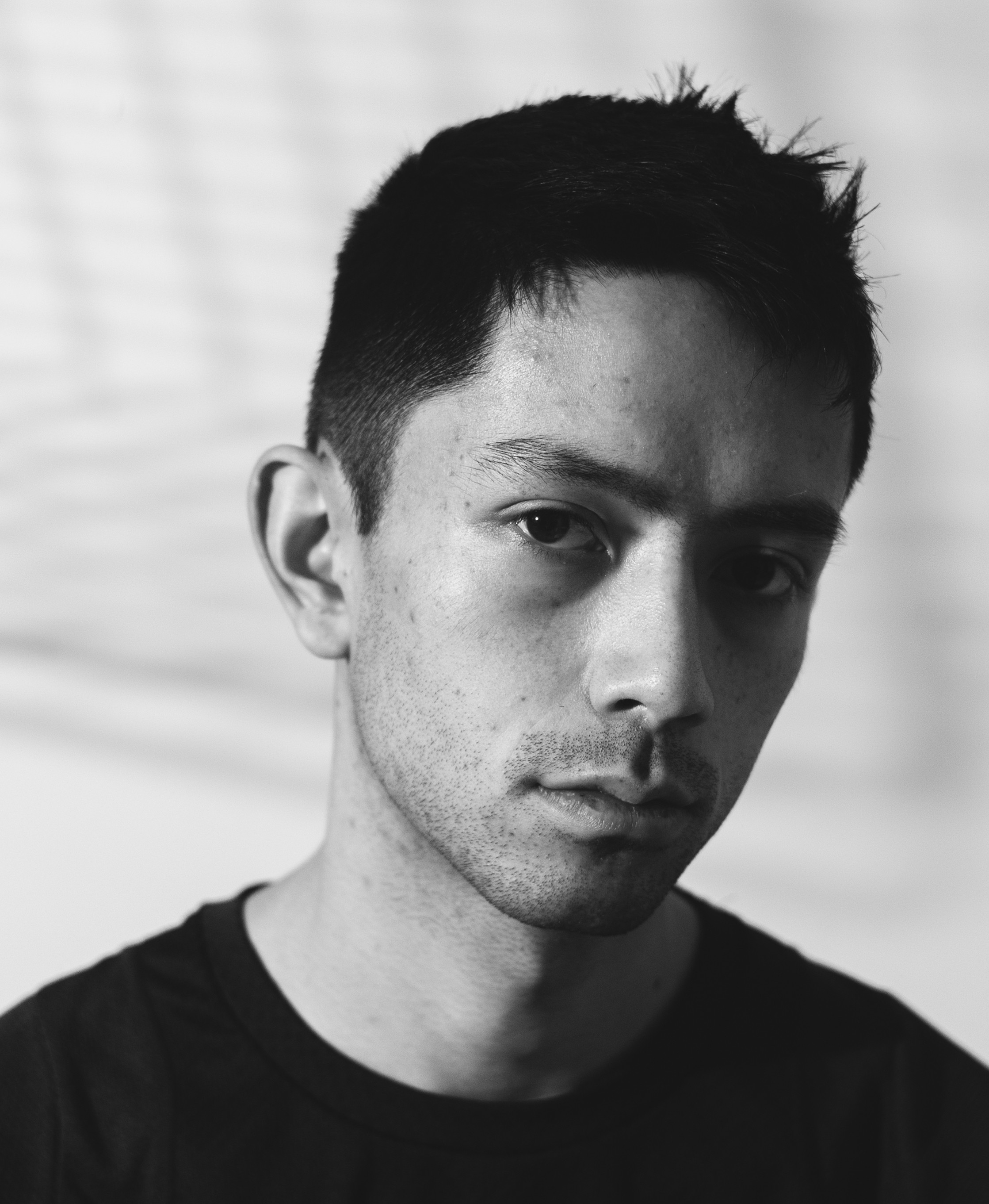 Black and white image of a man in a black shirt and short black hair gazing at the camera