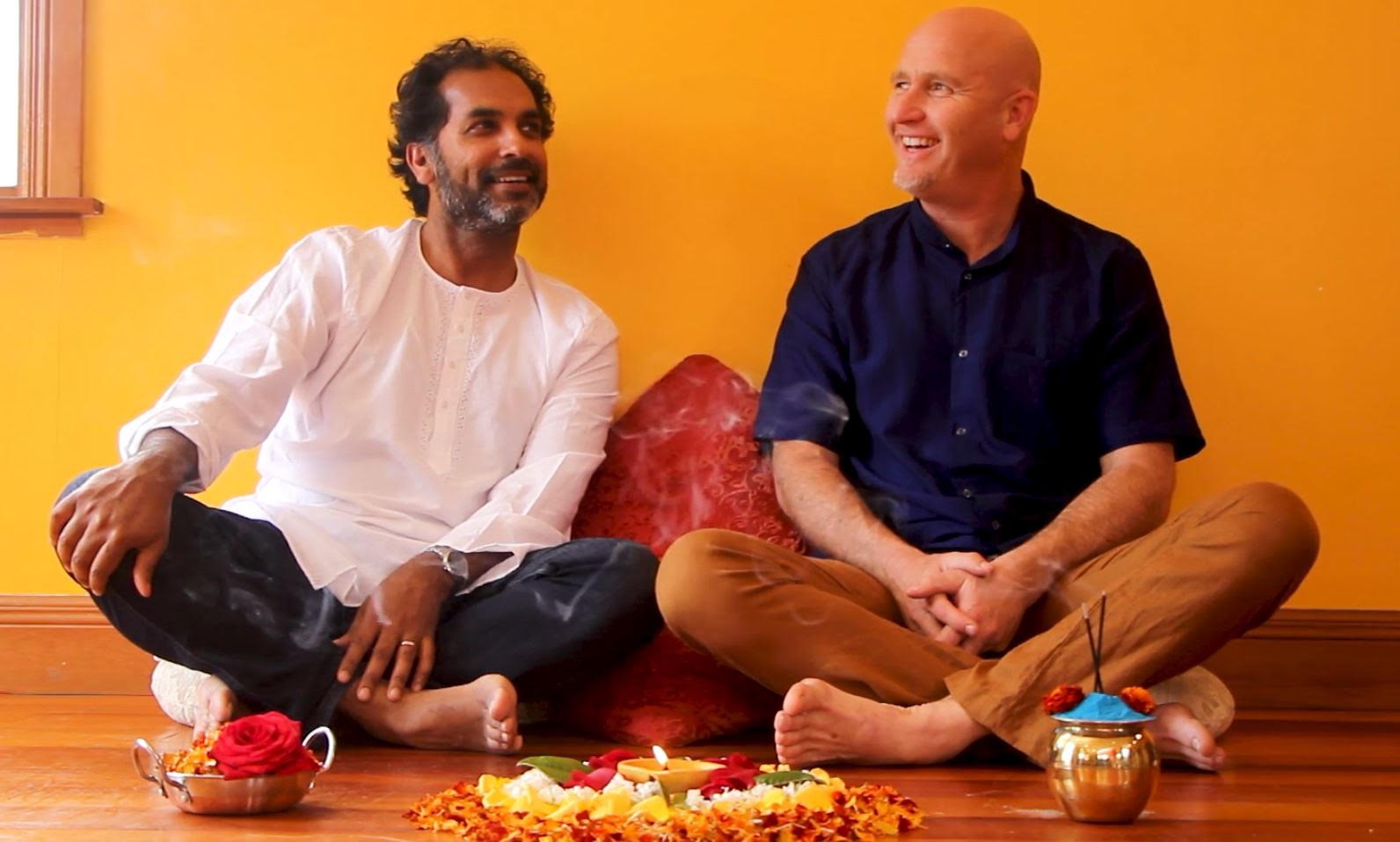 Two men sit cross legged in front of a yellow wall on a wooden floor. A candle surrounded by flowers is laid out in front of them, along with a bowl of flowers and some incense.