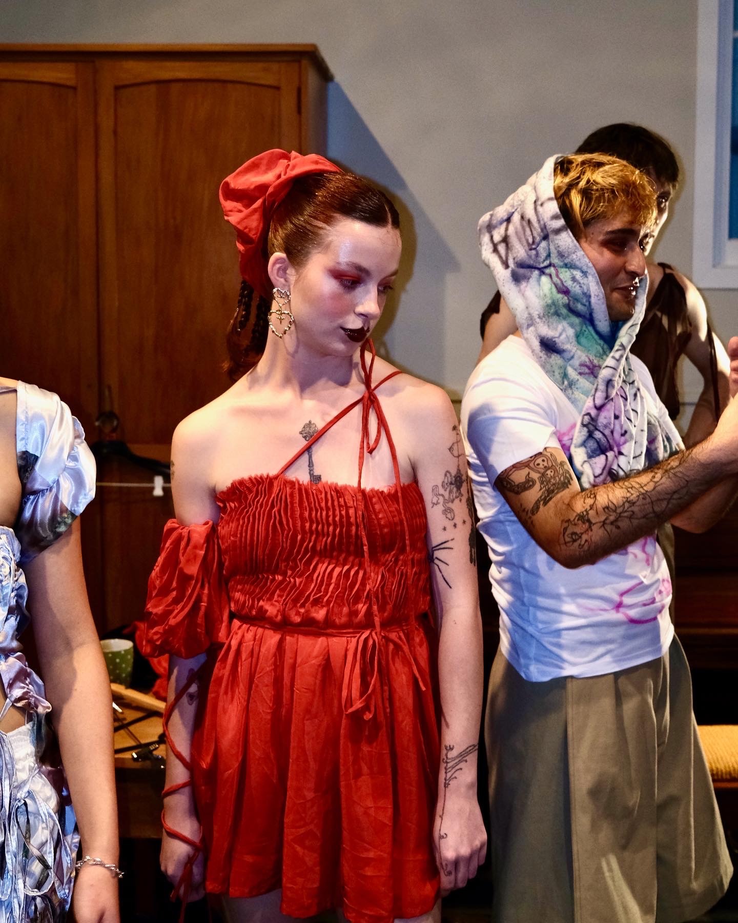 A model in a red dress with a ruched bodice, ties, and a matching scrunchie stands backstage at a fashion show