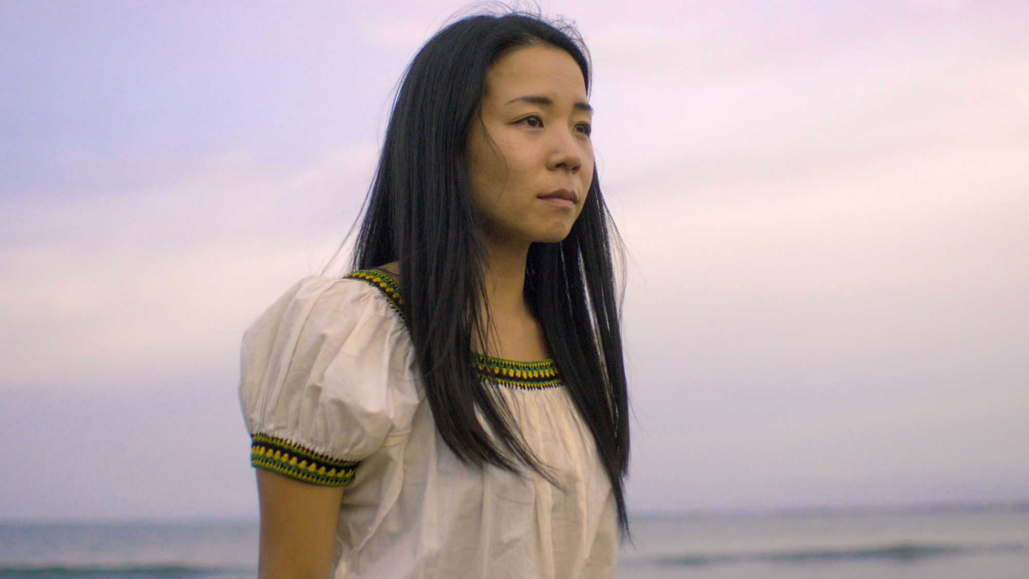 In a scene from ‘Kāinga’, a woman stands on a beach with the ocean behind her.