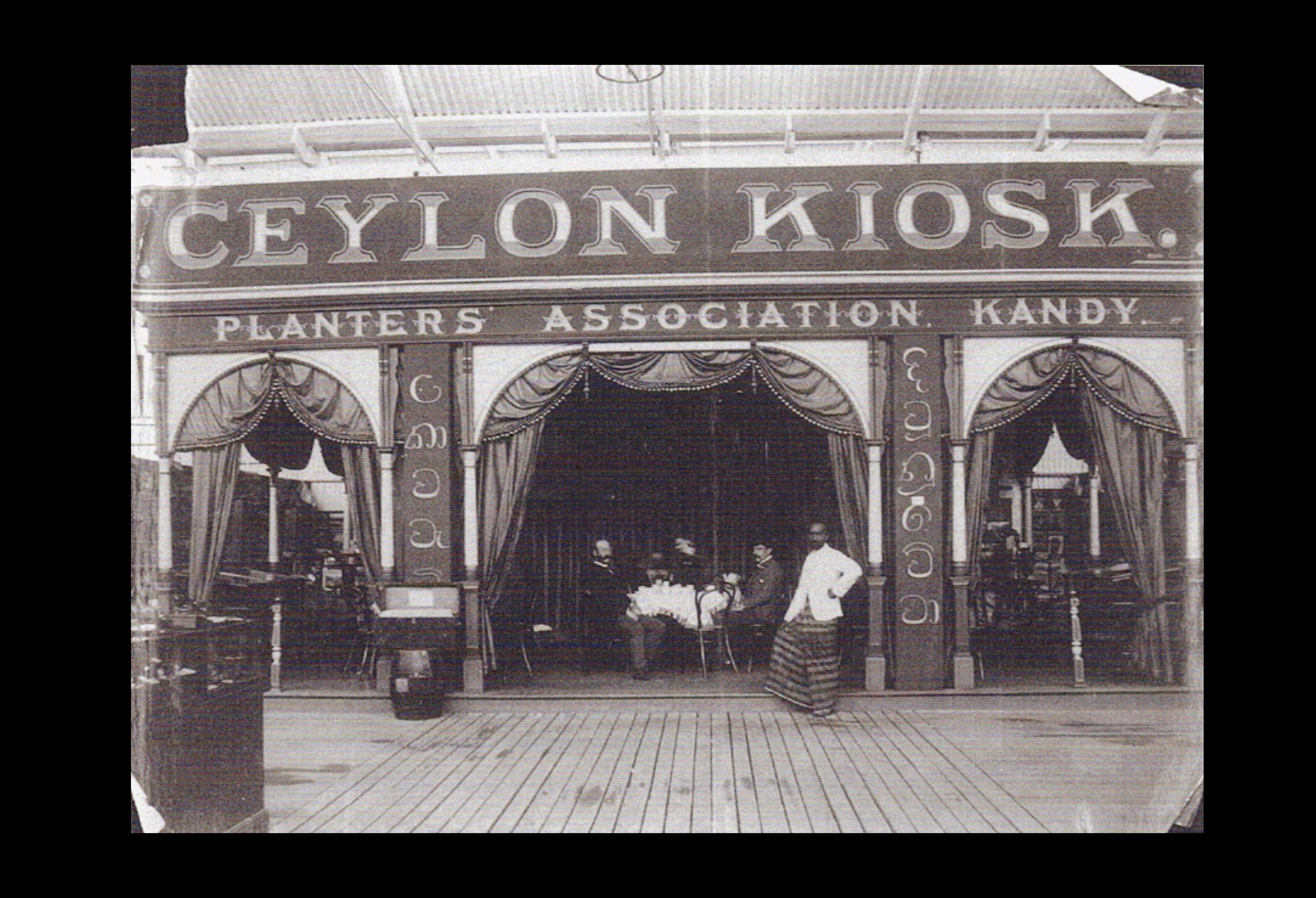 Black and white photograph of an elaborate kiosk with draped curtains and signage reading: Ceylon Kiosk - Planters Association Kandy.