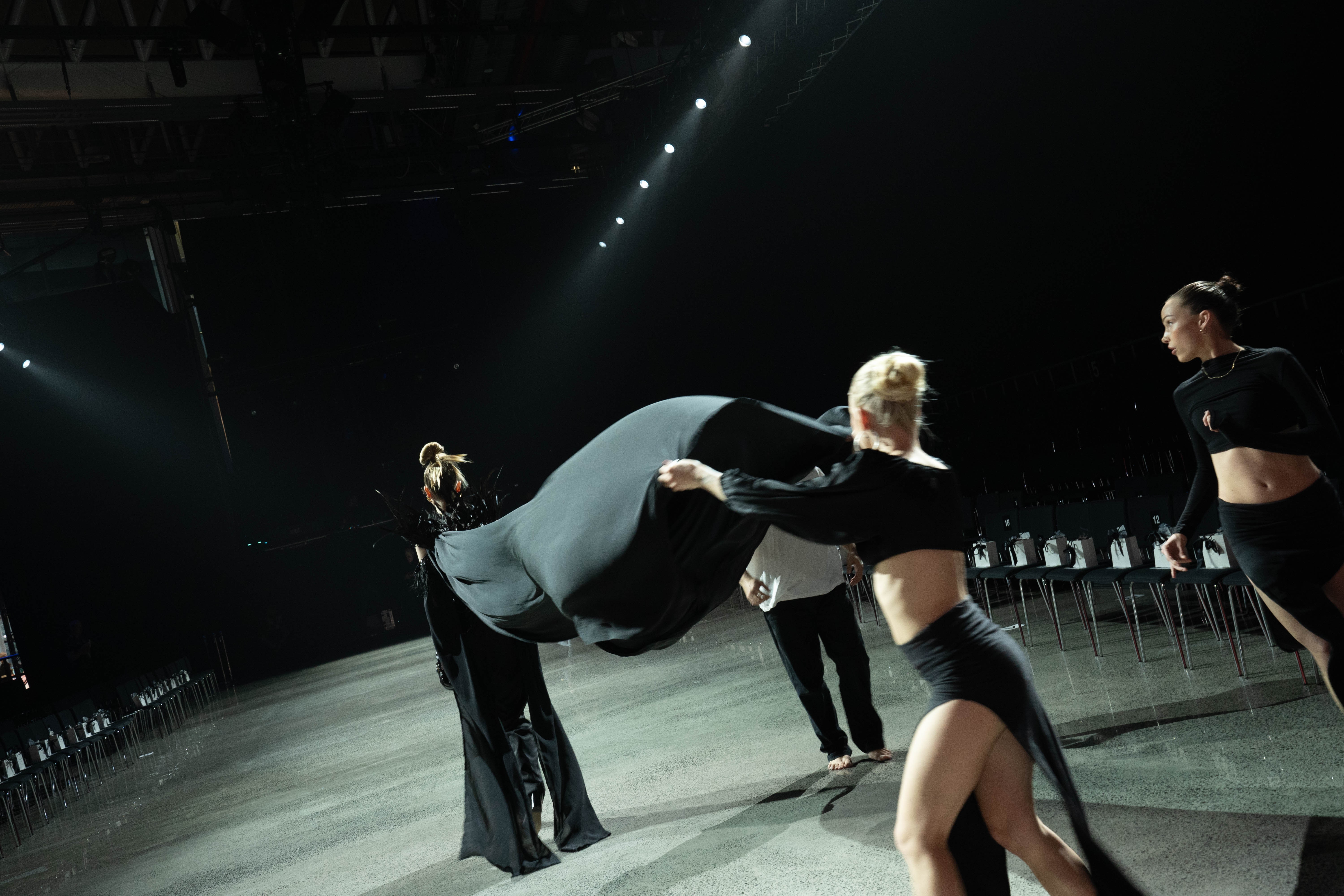 A woman with a long black train walks down a concrete runway while another model holds her train, which is billowing in the air