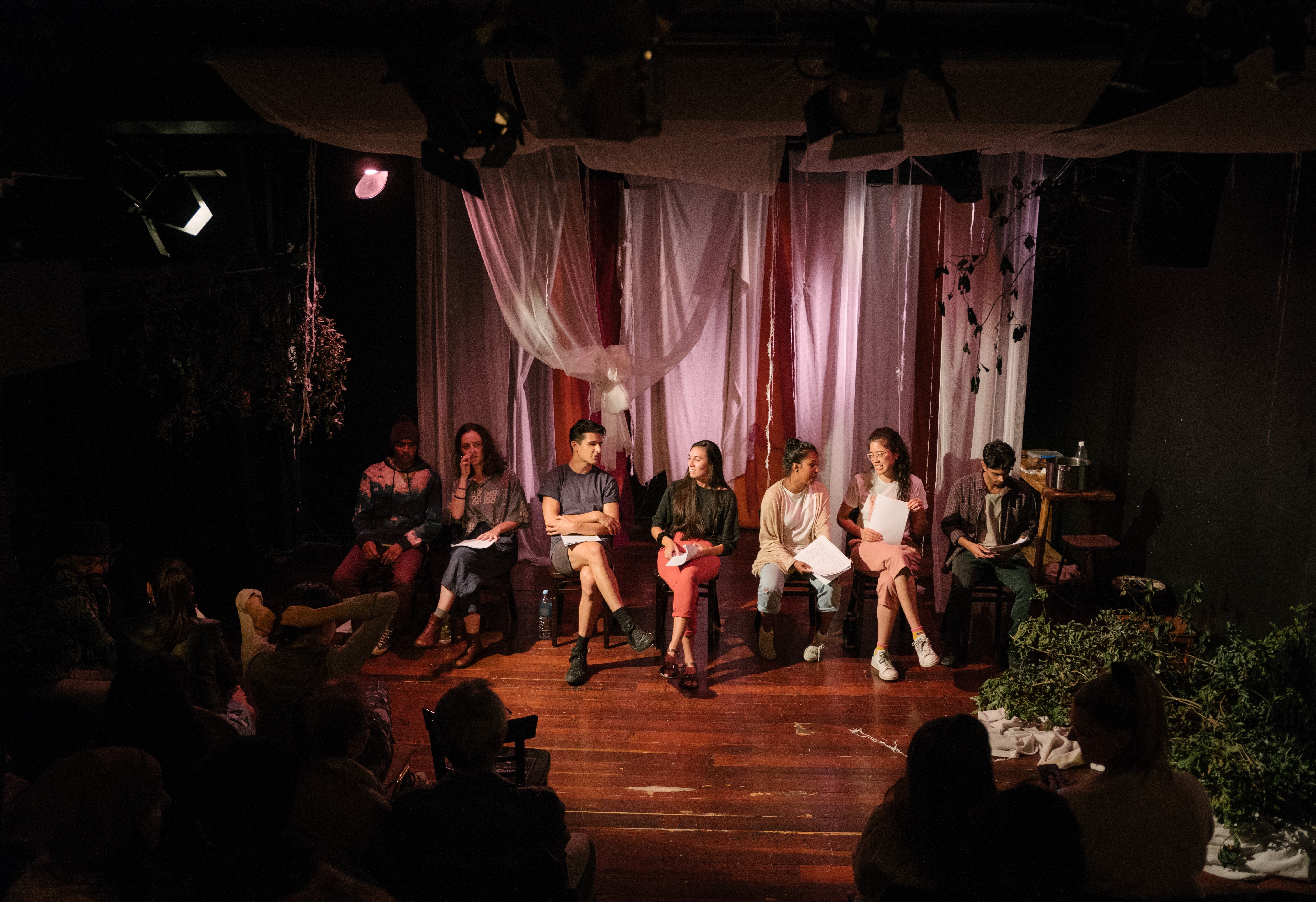 Seven actors sit on a line of chairs with scripts on their lap. Behind them pink mesh hangs romantically as stage dressing.