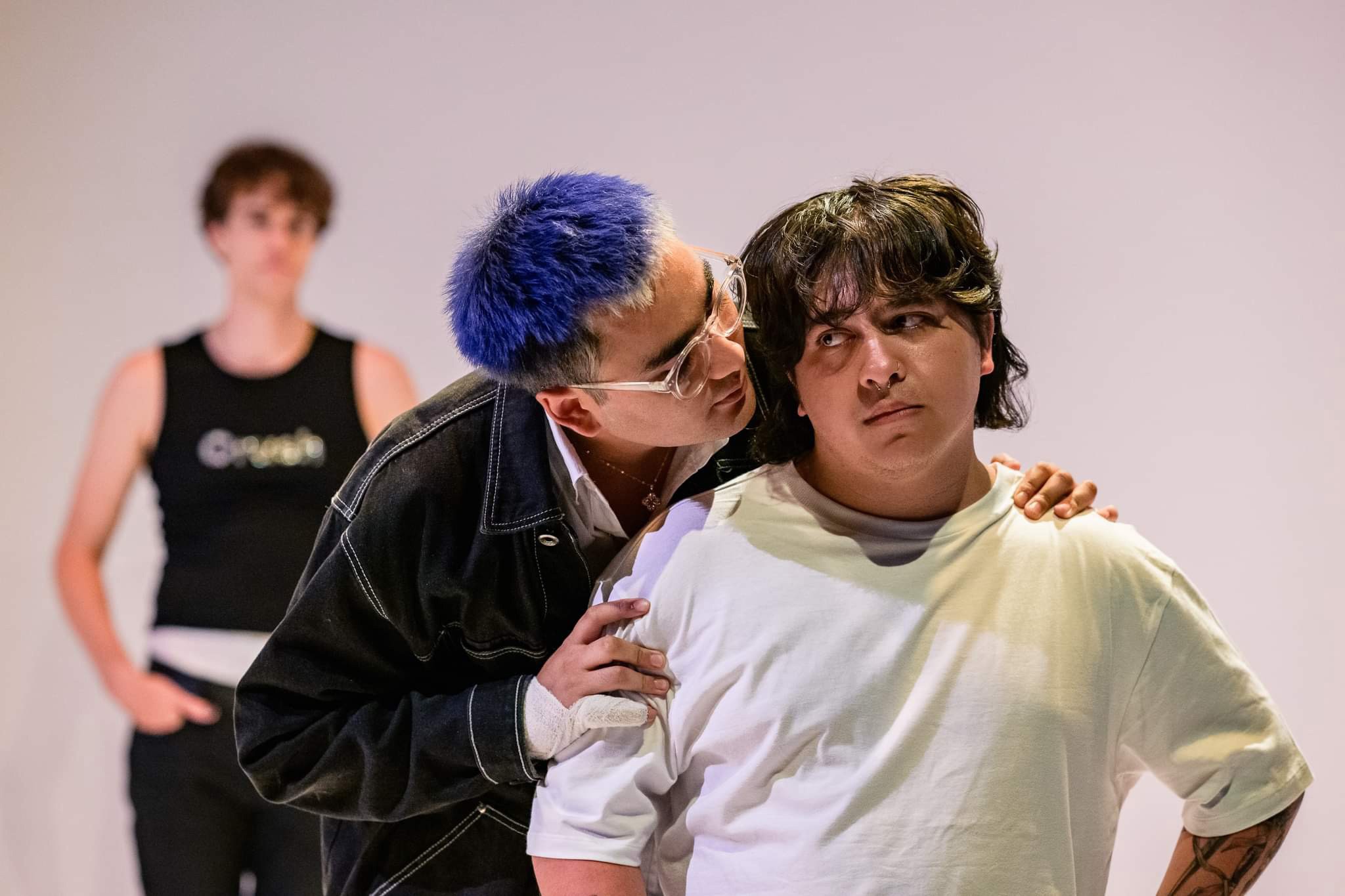 Person with unkempt hair and wearing a white shirt frowning while someone with blue hair wearing glasses and a black jacket is hugging them from the back