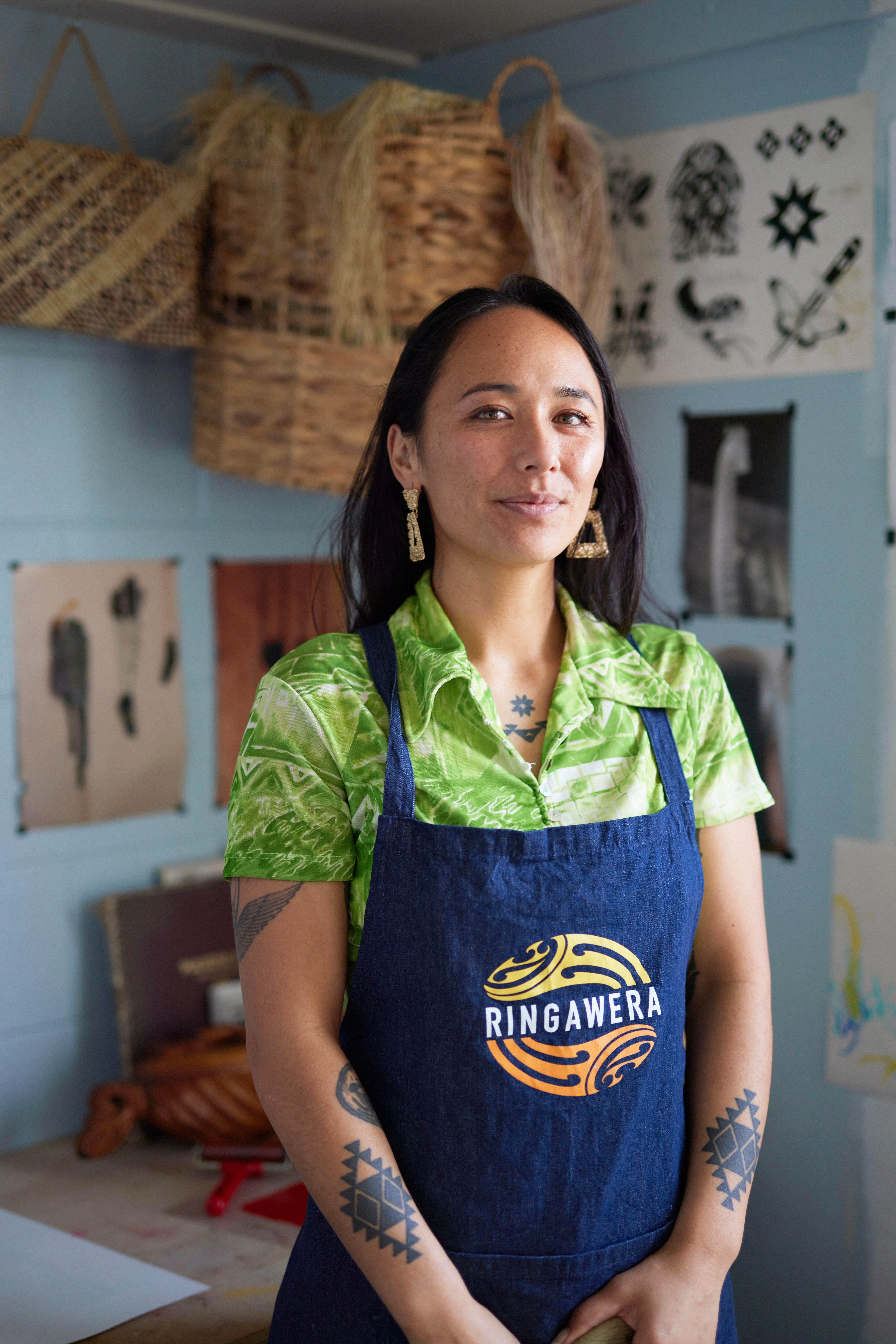 Emiko Sheehan stands in front of a wall covered with art. She wears a bright green shirt, long earrings, has tattoos on her arms, and an apron that says 'ringawera'