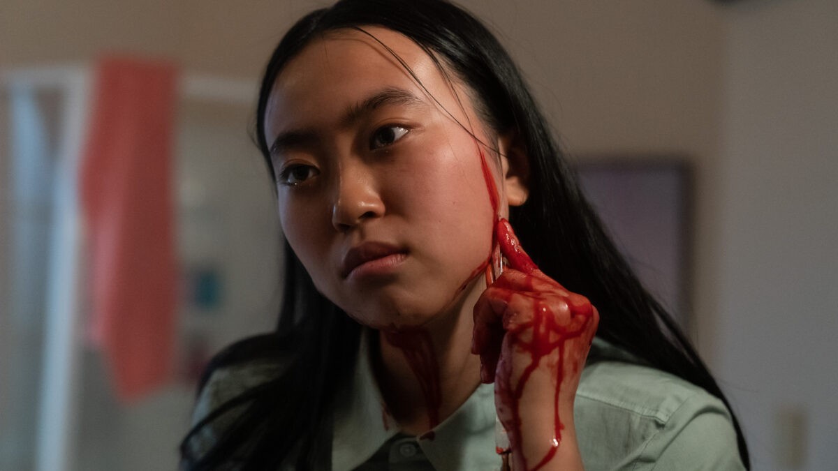 A movie still from a horror movie of an East Asian lady holding a scalpel to her face and drawing blood