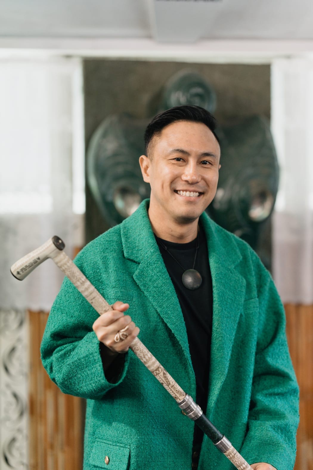 Chris Tse with short black hair and dimples, smiles at the camera. He is wearing a green coat and pounamu and holding a carved tokotoko.
