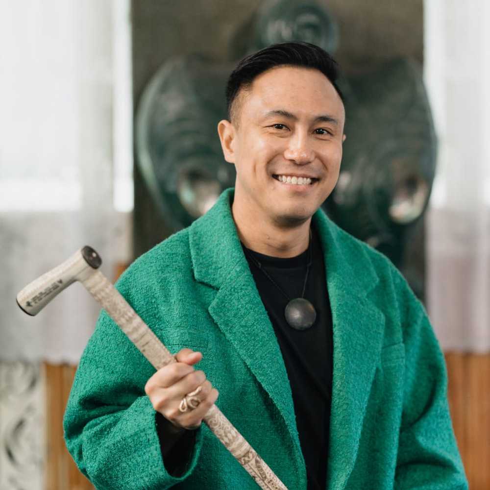 Chris Tse with short black hair and dimples, smiles at the camera. He is wearing a green coat and pounamu and holding a carved tokotoko.