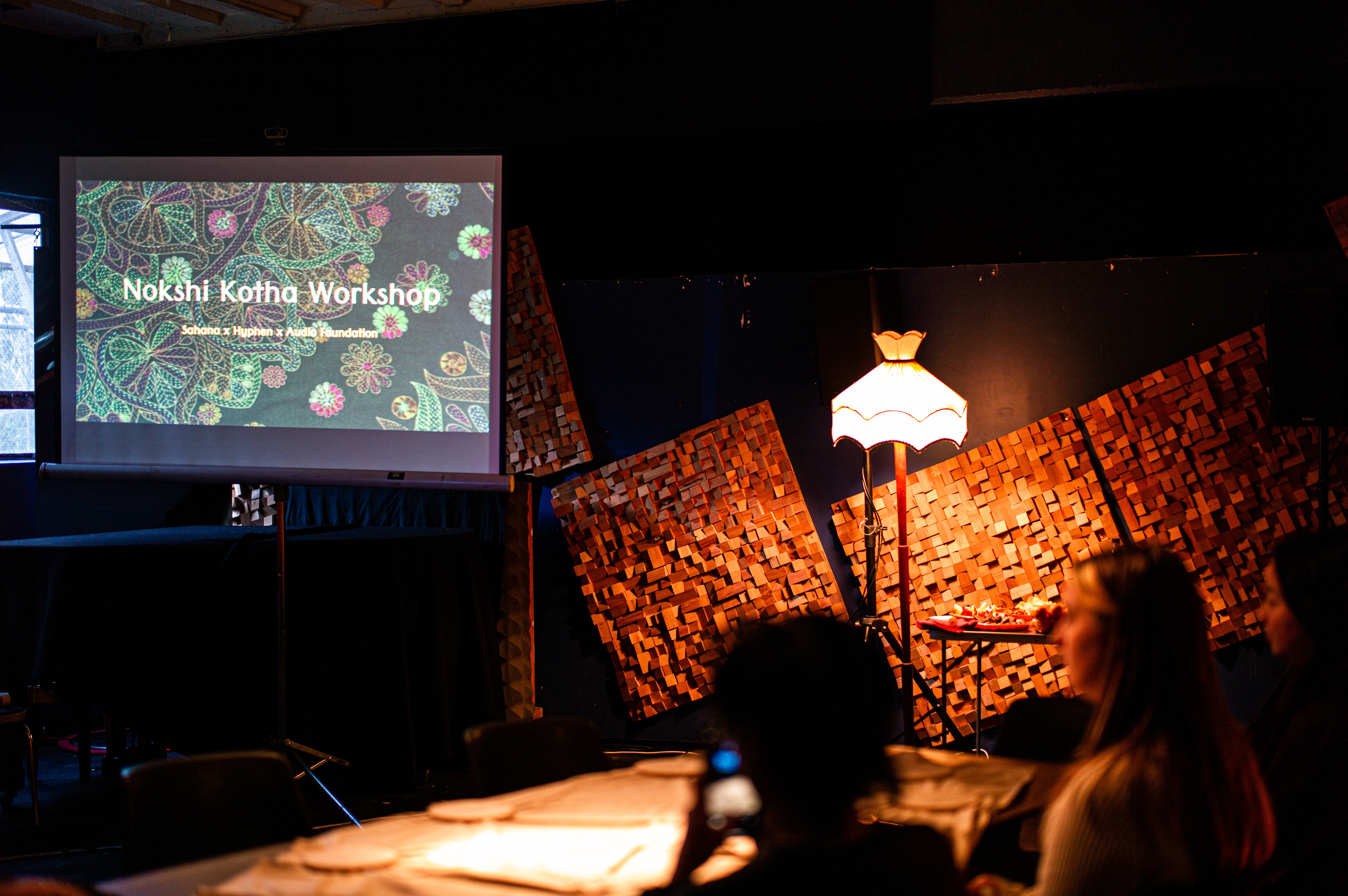 A darkened room lit by a single lamp with a projection showing the words Nokshi Kotha Workshop, with two participants sitting at a table facing the screen. 