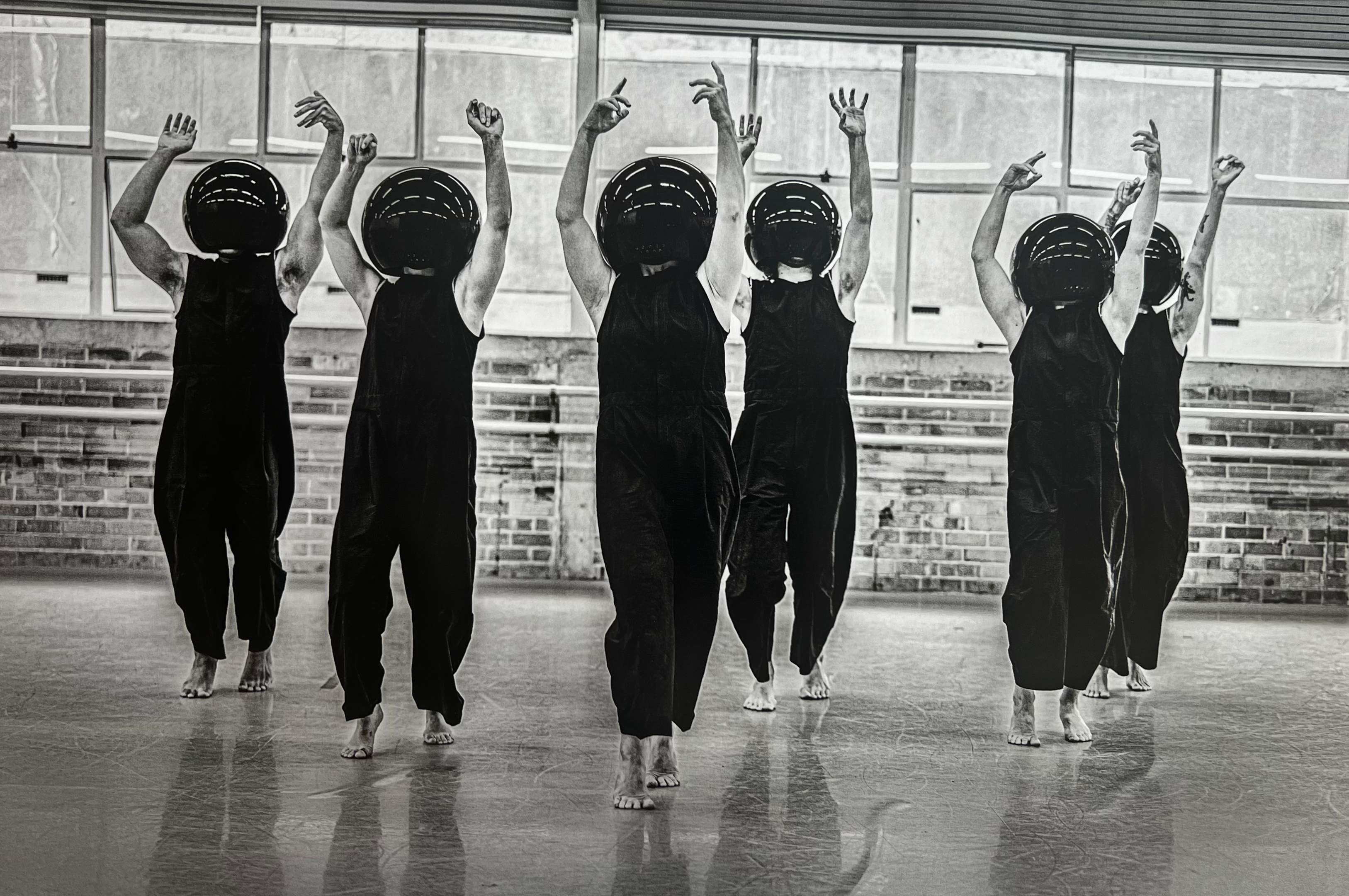 A group of dancers in formation standing on their toes wearing identical round black helmets that obscure their faces