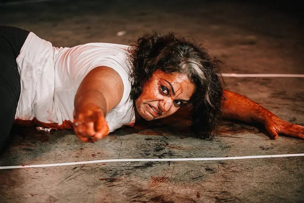 A woman splattered in blood lying on a concrete stage floor pointing angrily off-camera