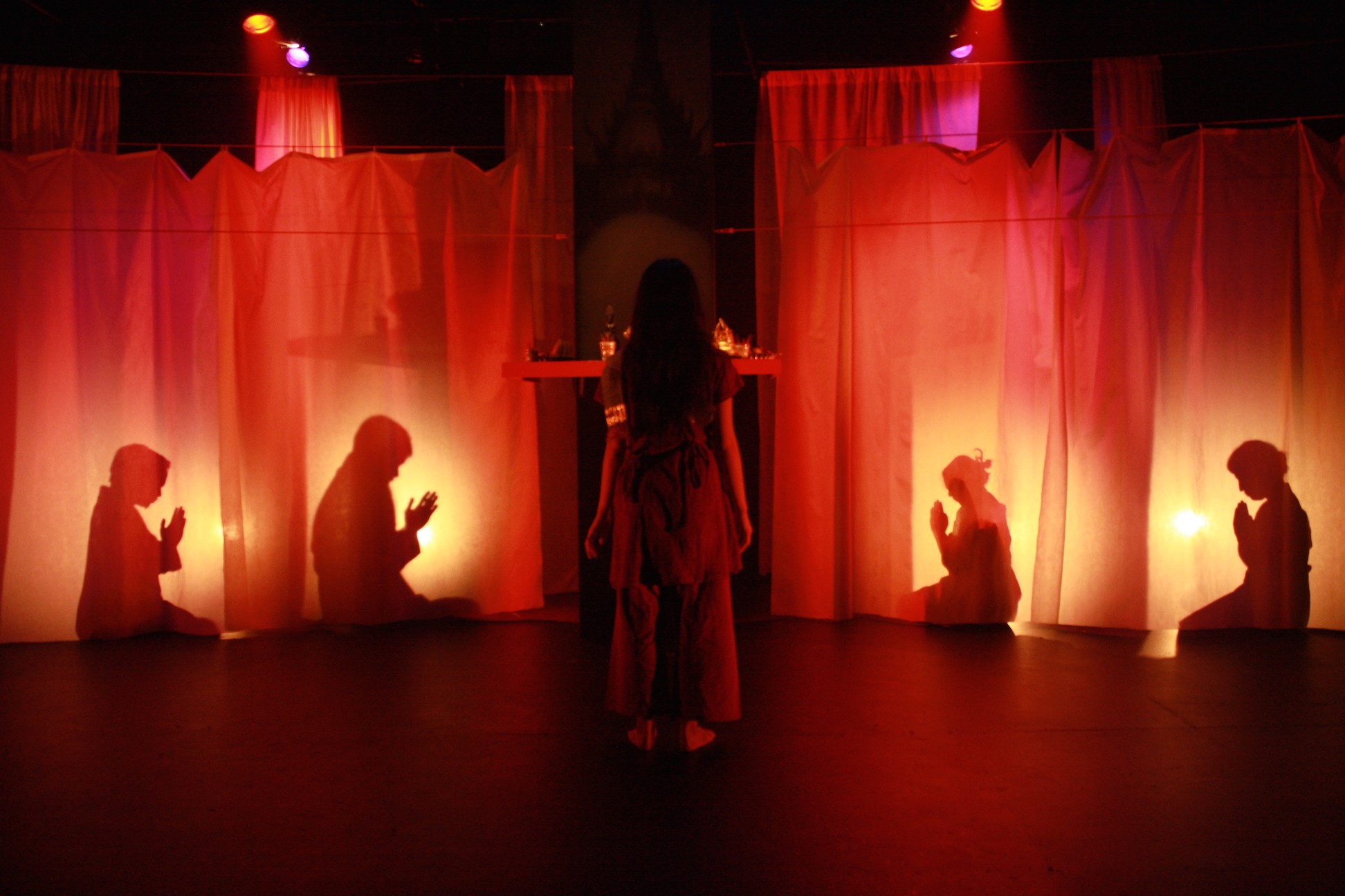 A woman faces the back of stage where praying actors are silhouetted behind gauzy curtains.