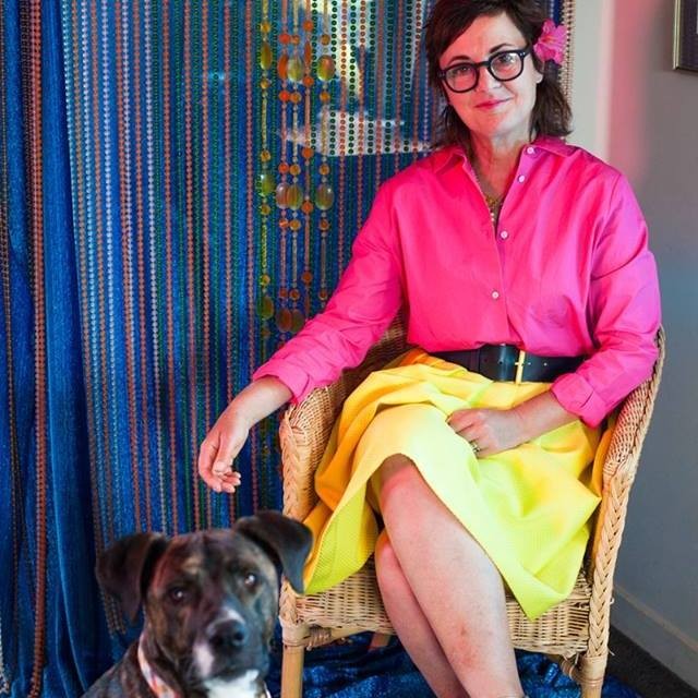 Laurie Foon sits in front of a beaded curtain and next to a dog. She wears glasses, and a bright pink and yellow outfit and has a pink flower in her hair.