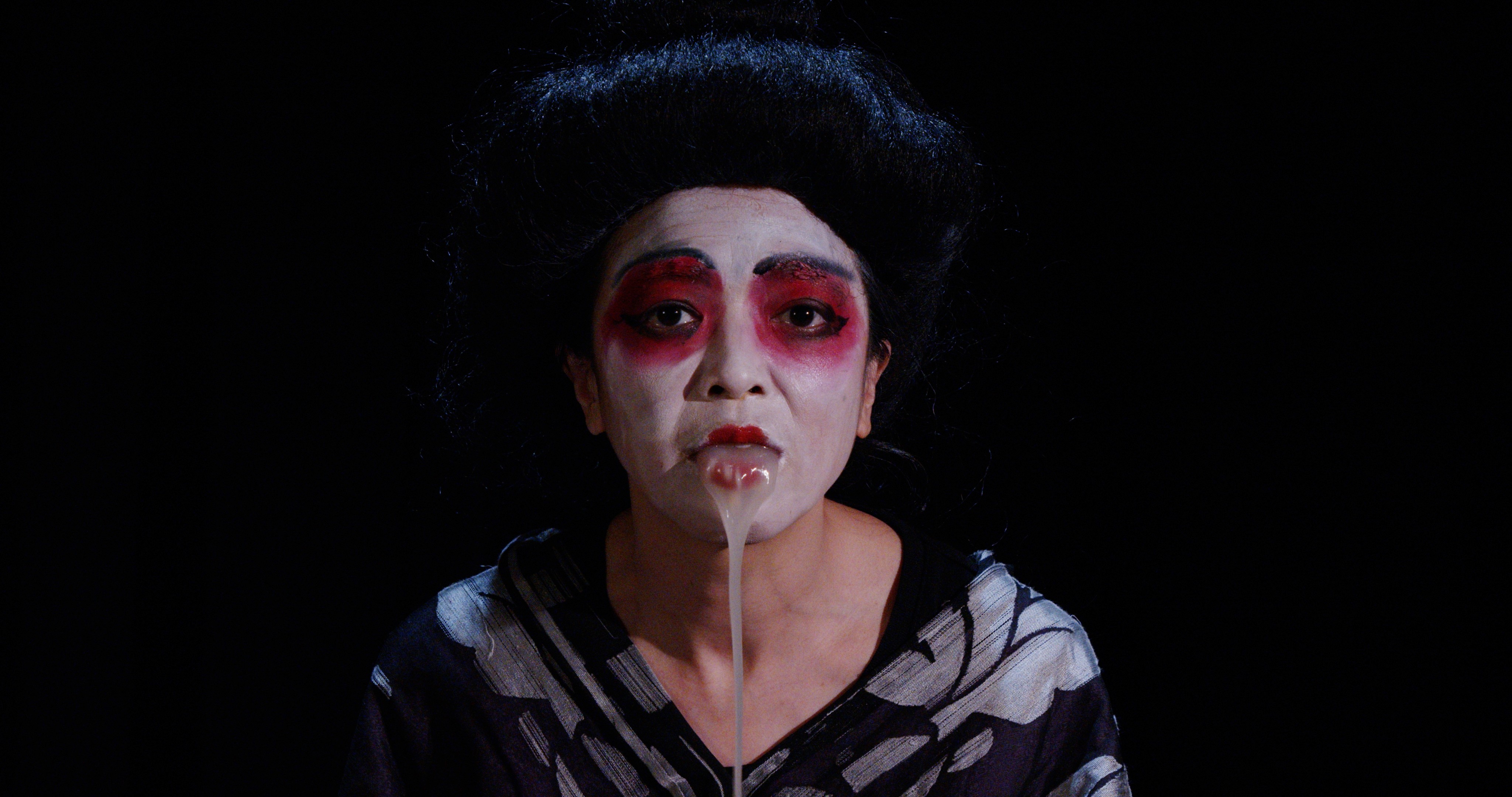 Image of a hand holding a small rectangular mirror. Reflected in the mirror is a woman whose face is painted white with red lipstick and bright red eyeshadow encircling her eyes.