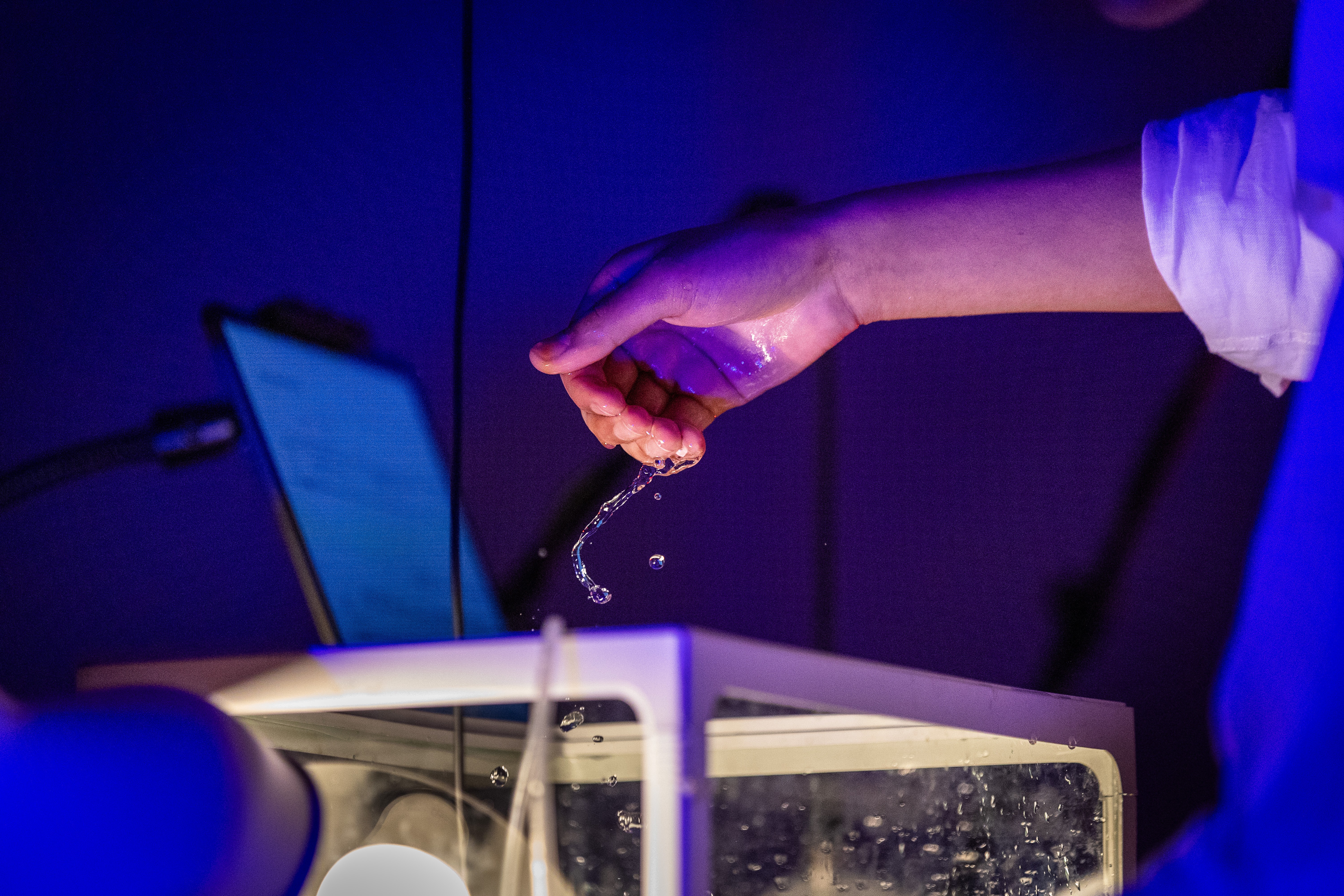 Photo of a performer's hand cupping dripping water above a tank, in a blue and purple atmosphere