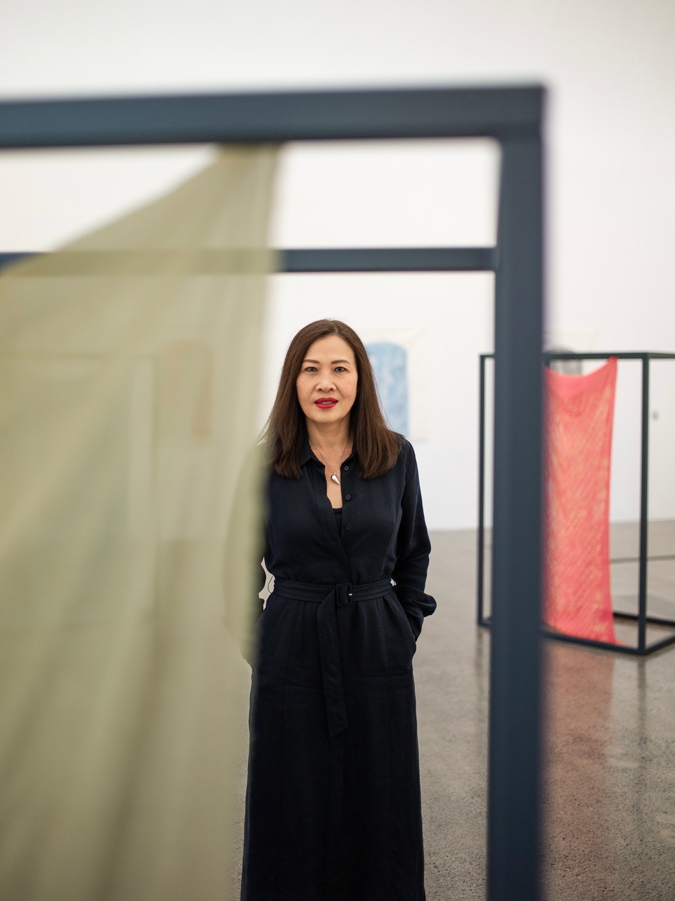 Rozana has long hair and is wearing a long black dress. She stands between artworks with gauzy fabric suspended within frames.