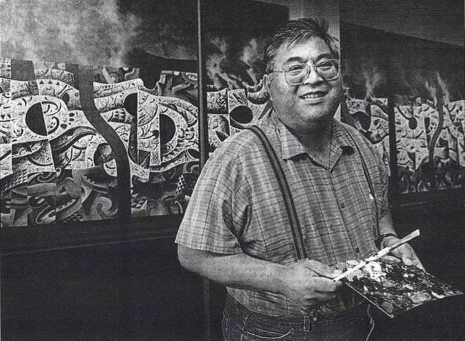 A smiling man wearing glasses wears a short-sleeved checked shirt with suspenders. He holds a paint palette and brush and is standing in front of a painting.