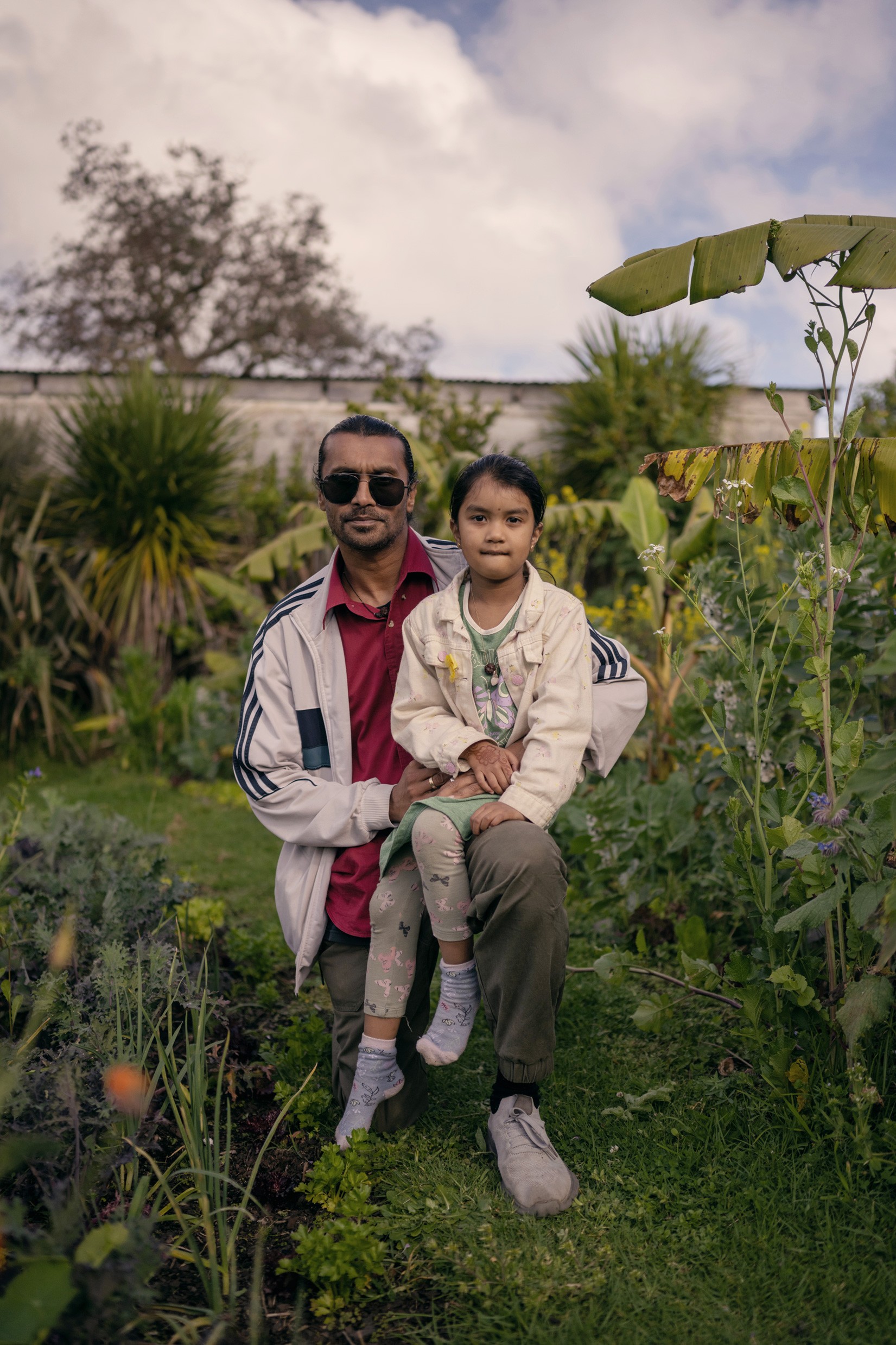 Person with sunnies and daughter sitting on their knee, in front of a lush garden