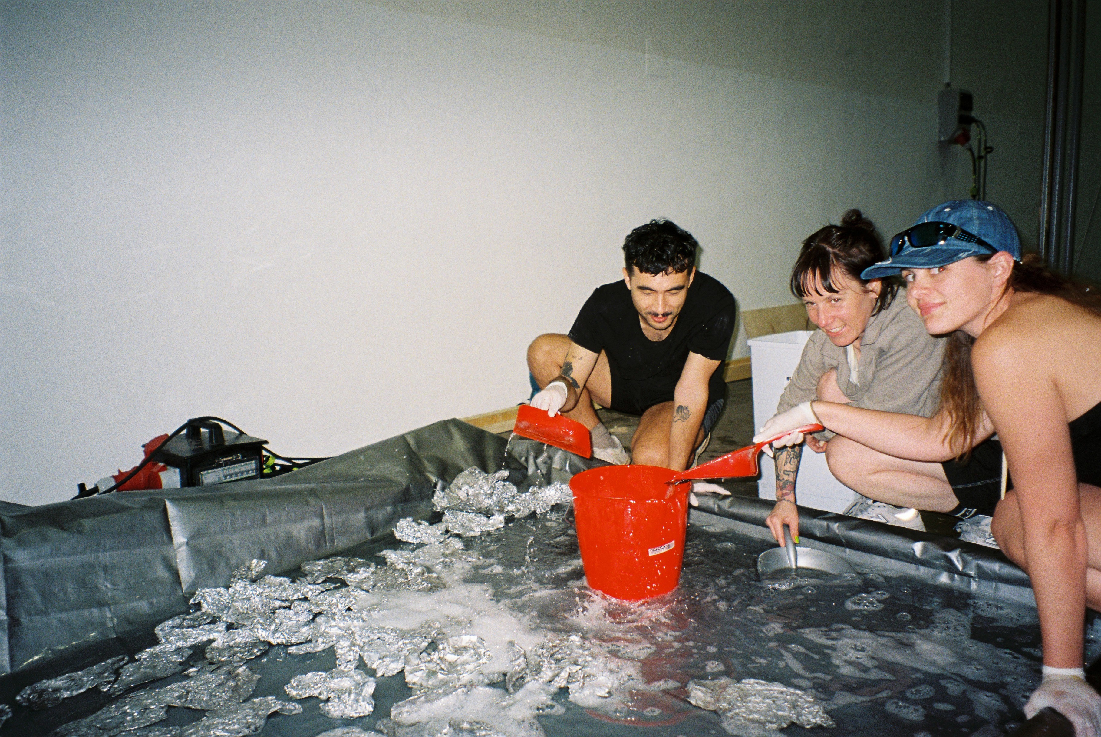 Three people scooping water out of an inflatable pool. The pool also has pieces of aluminium foil in it.
