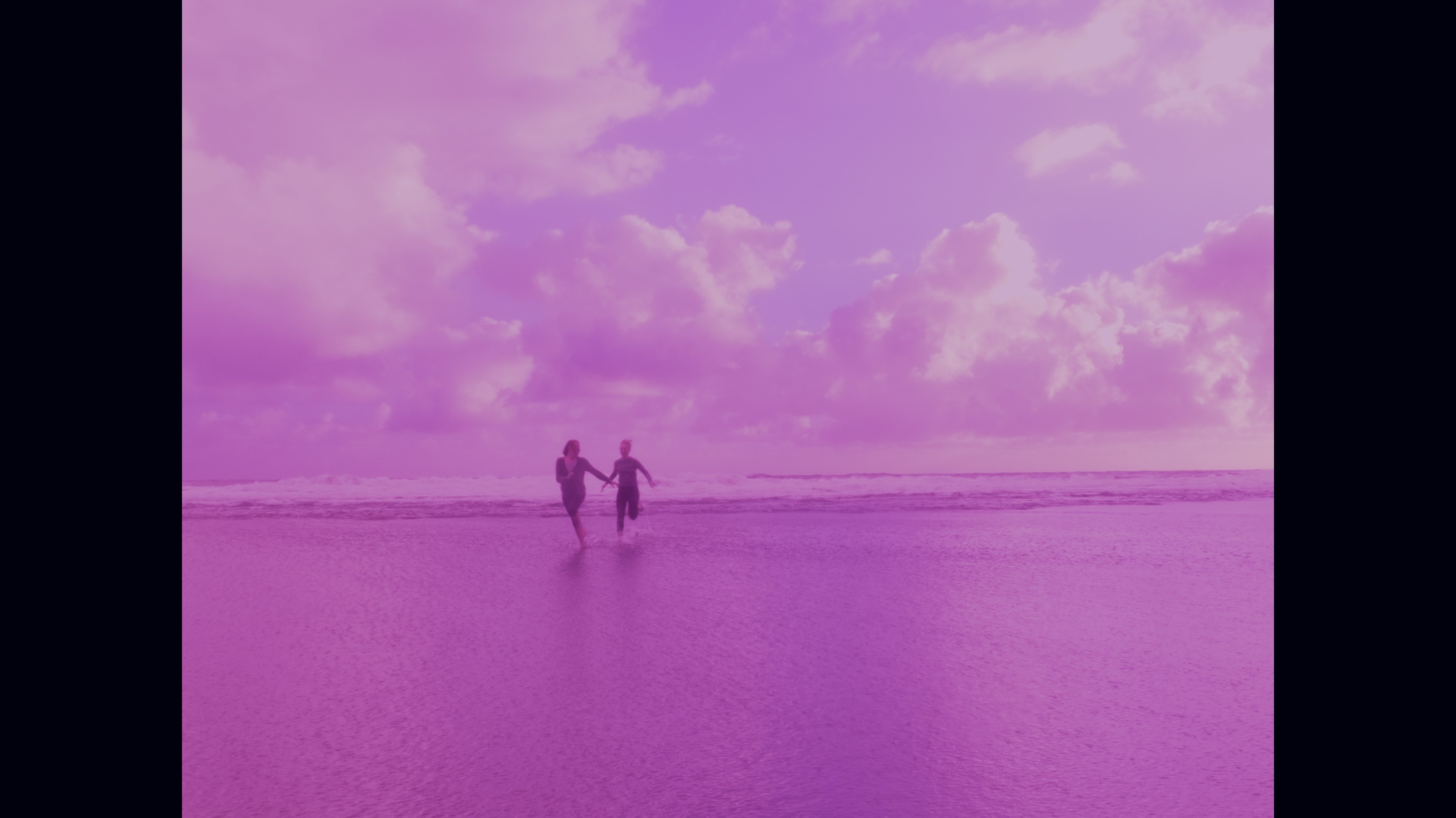 In a scene from the film "The Night That Holds You", two women in the distance, holding hands, run along a beach.