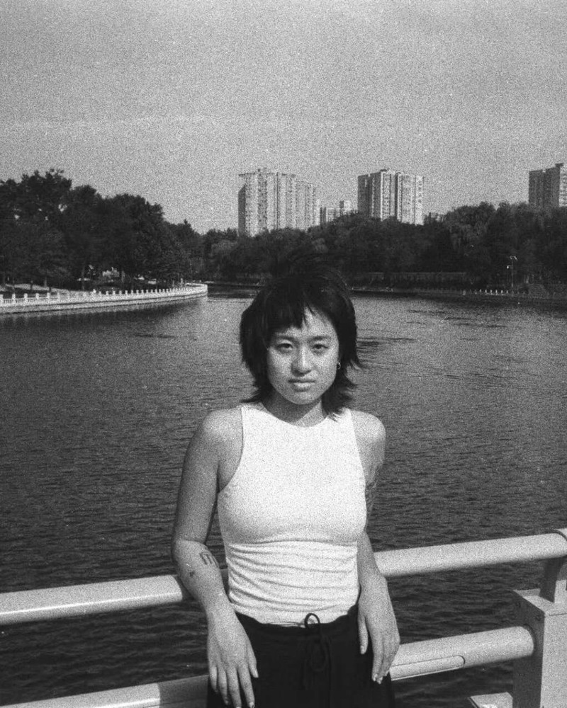 Jing is wearing a white singlet and has short shaggy hair. They are posing on a bridge in front of a river.