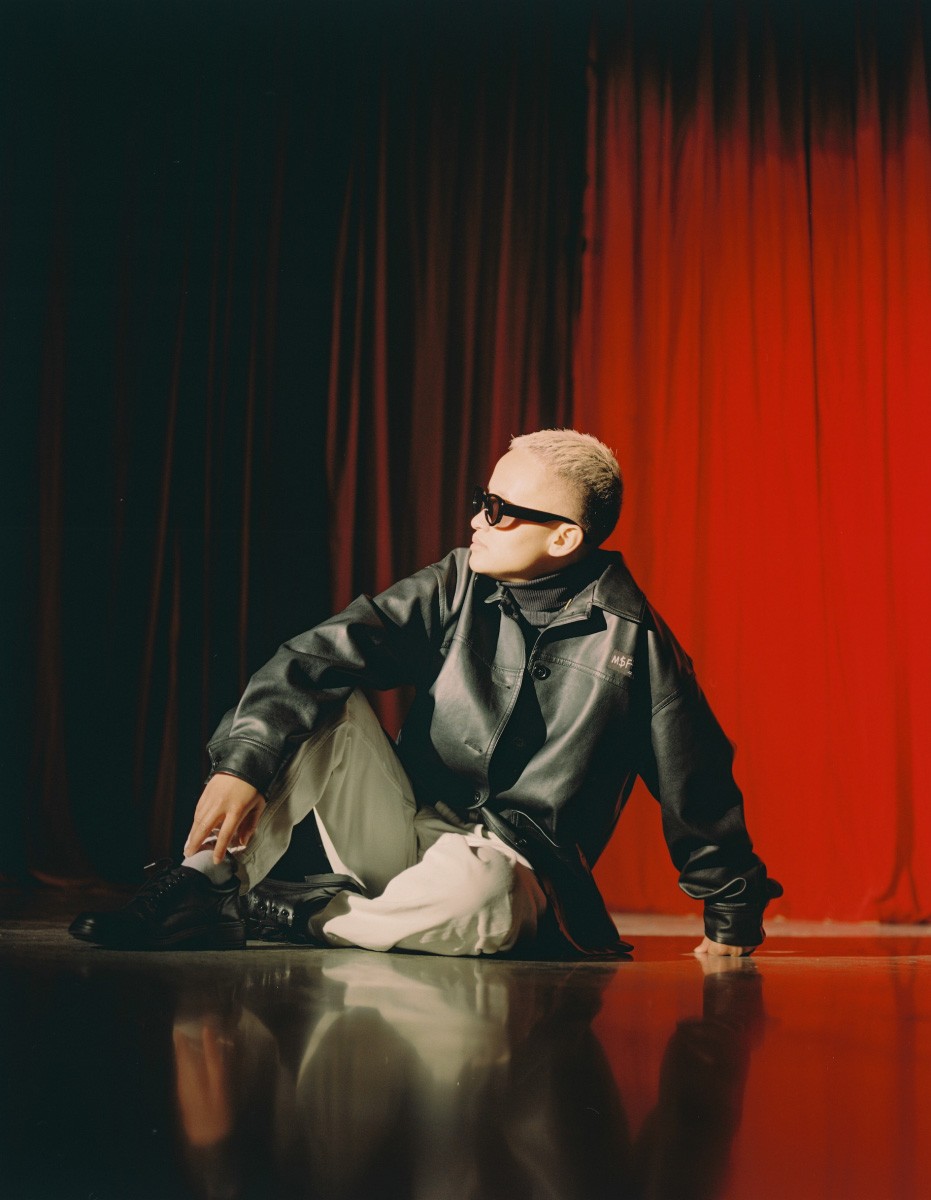 Filipino person sitting on a stage with red curtains, looking to the side wearing dark sunnies, a black leather jacket and khaki pants