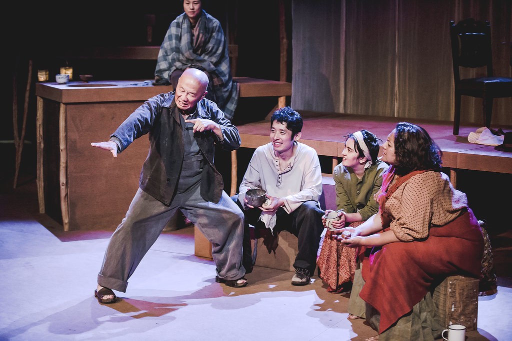 Actors on stage huddle around a man striking a pose as he tells a story.