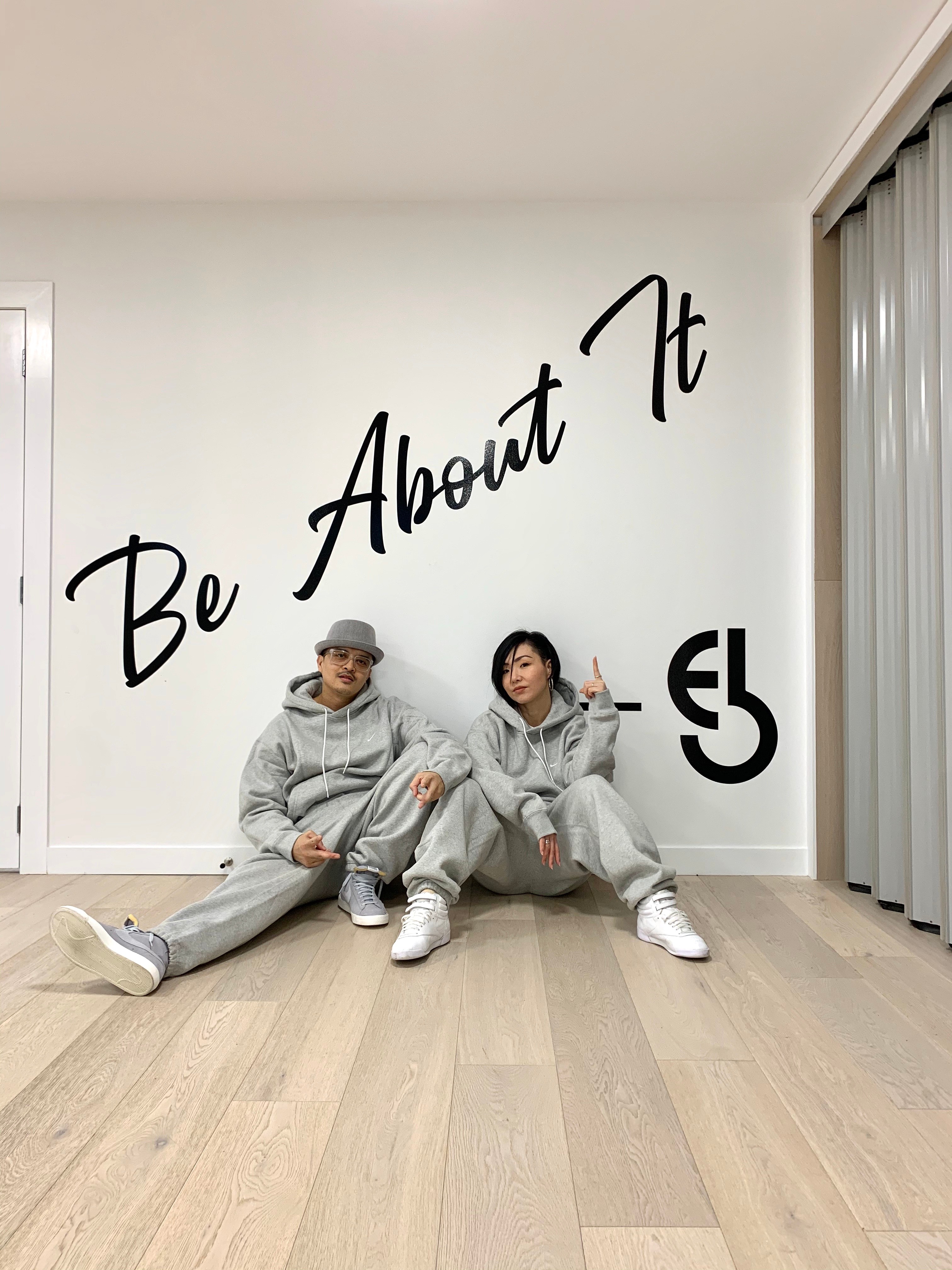 A man and woman in matching grey tracksuits sit on the ground posing in front of a wall that says 'Be About It'