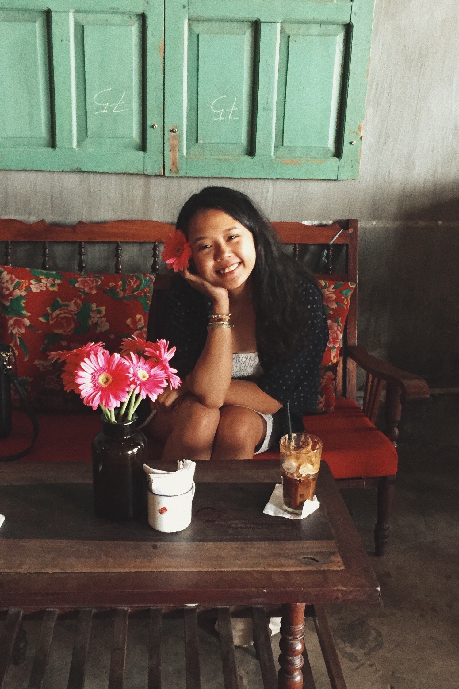 A smiling Wai Ching Chan has a pink hibiscus flower in her long dark wavy hair and is sitting next to a bunch of flowers.