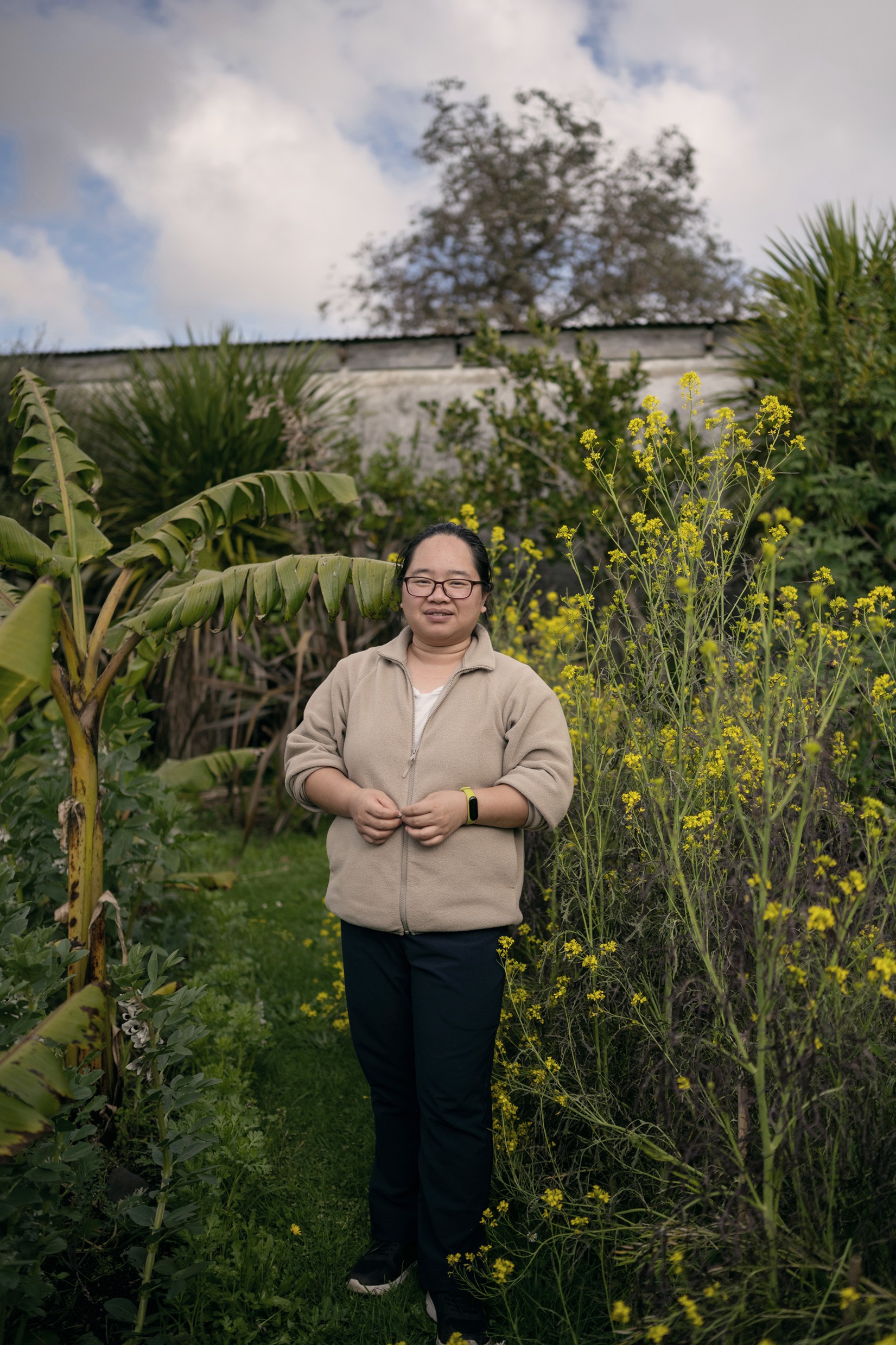 A person of Asian decent in a beige sweater standing in front of a lush garden