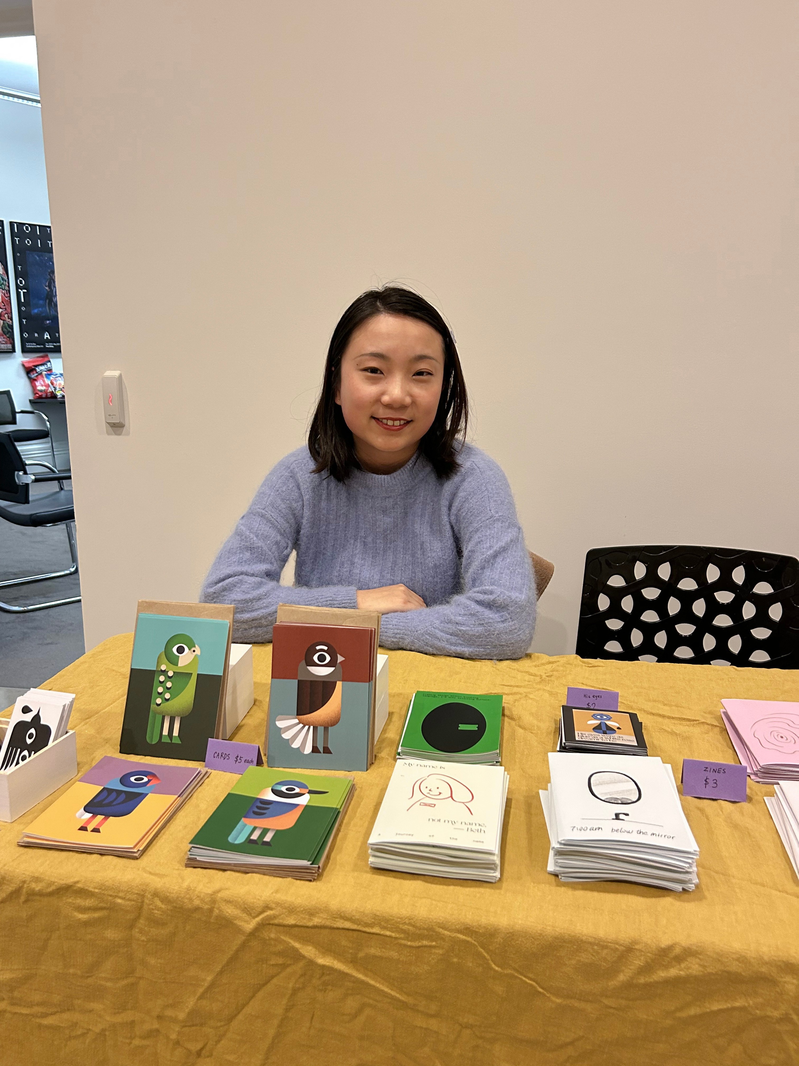 A person smiling sitting behind a zine table with various prints and zines displayed. 