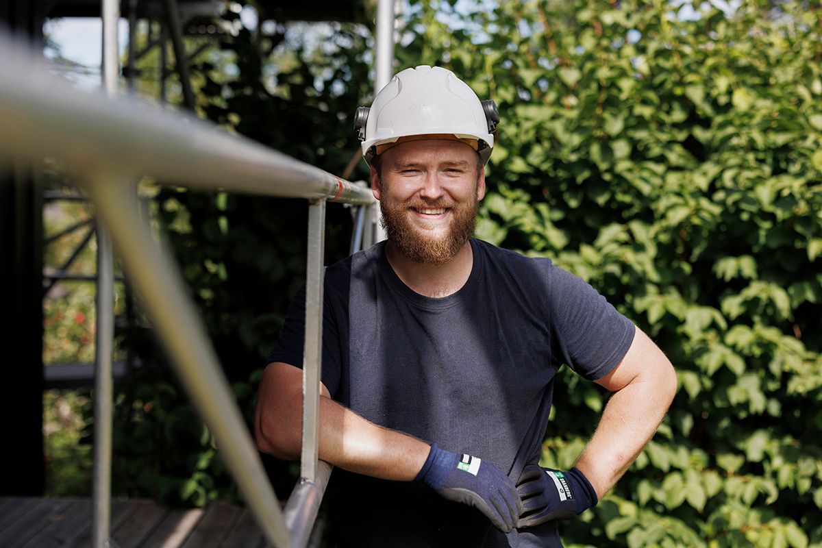 Tømrer med hjelm smilende ved stillaset under et takprosjekt.