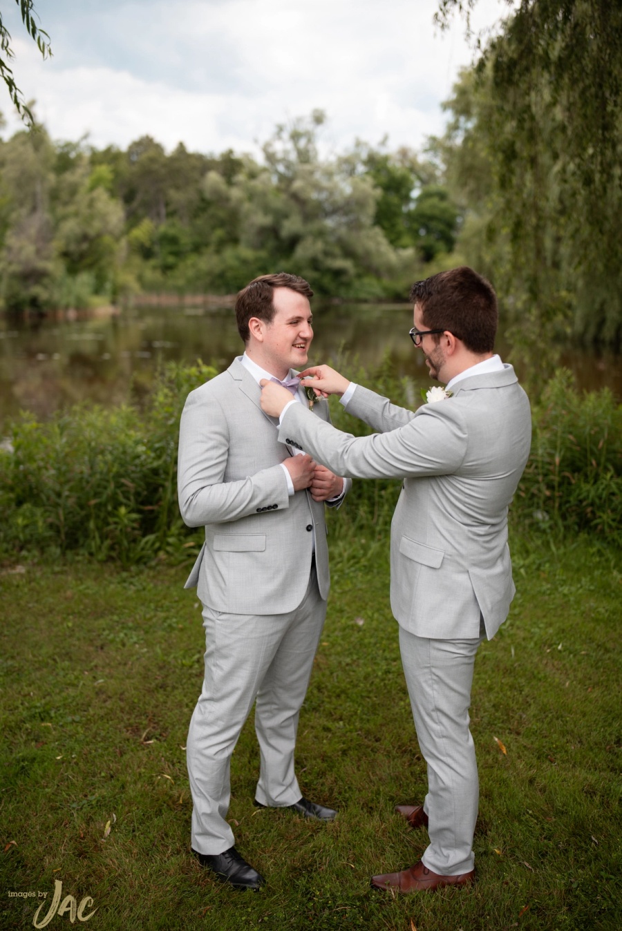 A groomsman straightens the groom's bow tie on a lawn beside the water