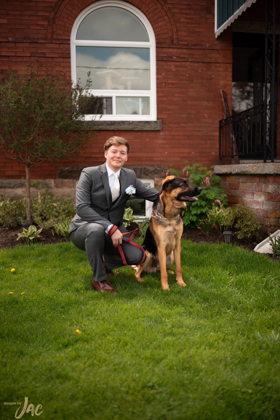 A groom kneels on the grass beside his dog outside a red brick house