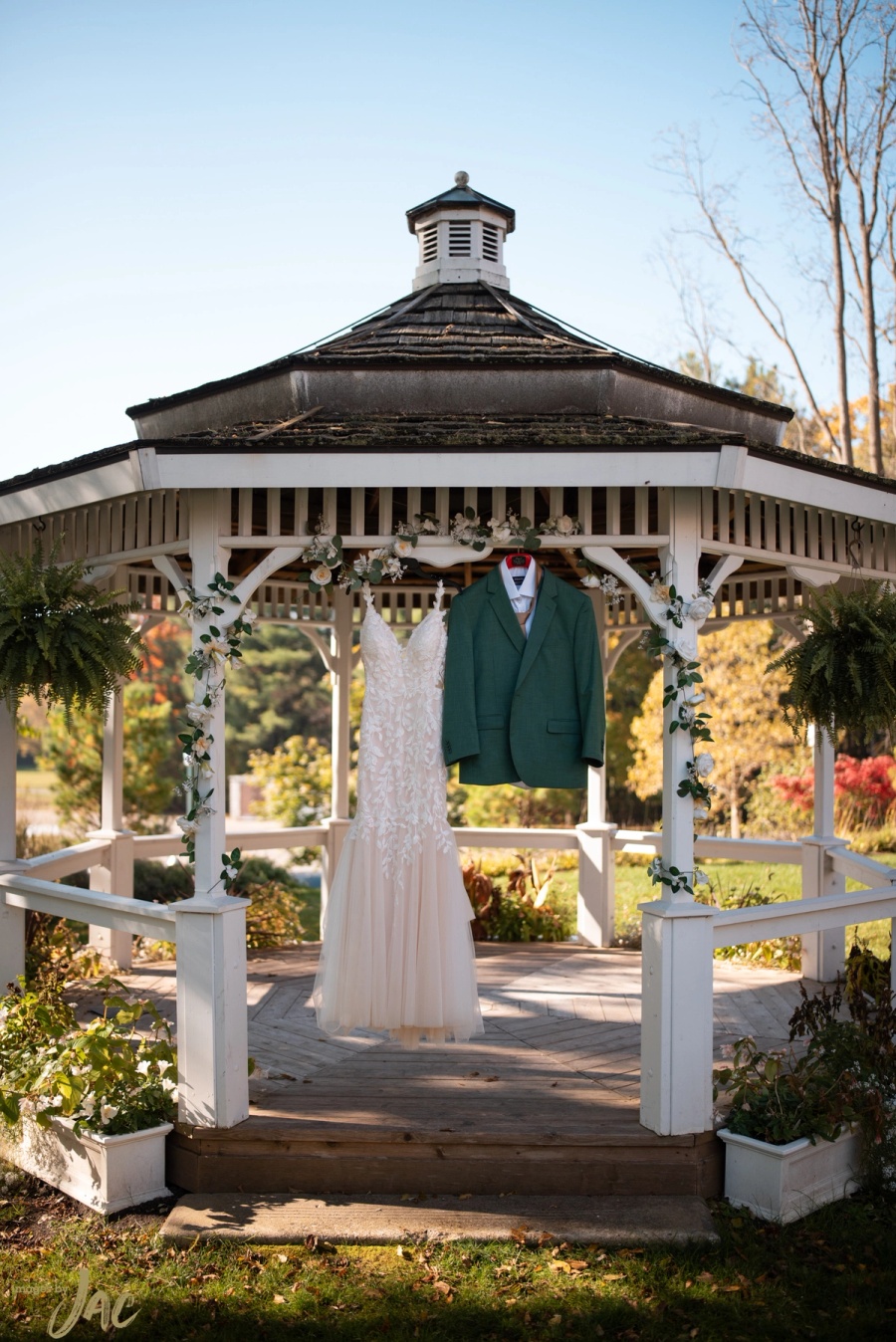 A wedding gown and the groom's green jacket hanging in a white gazebo