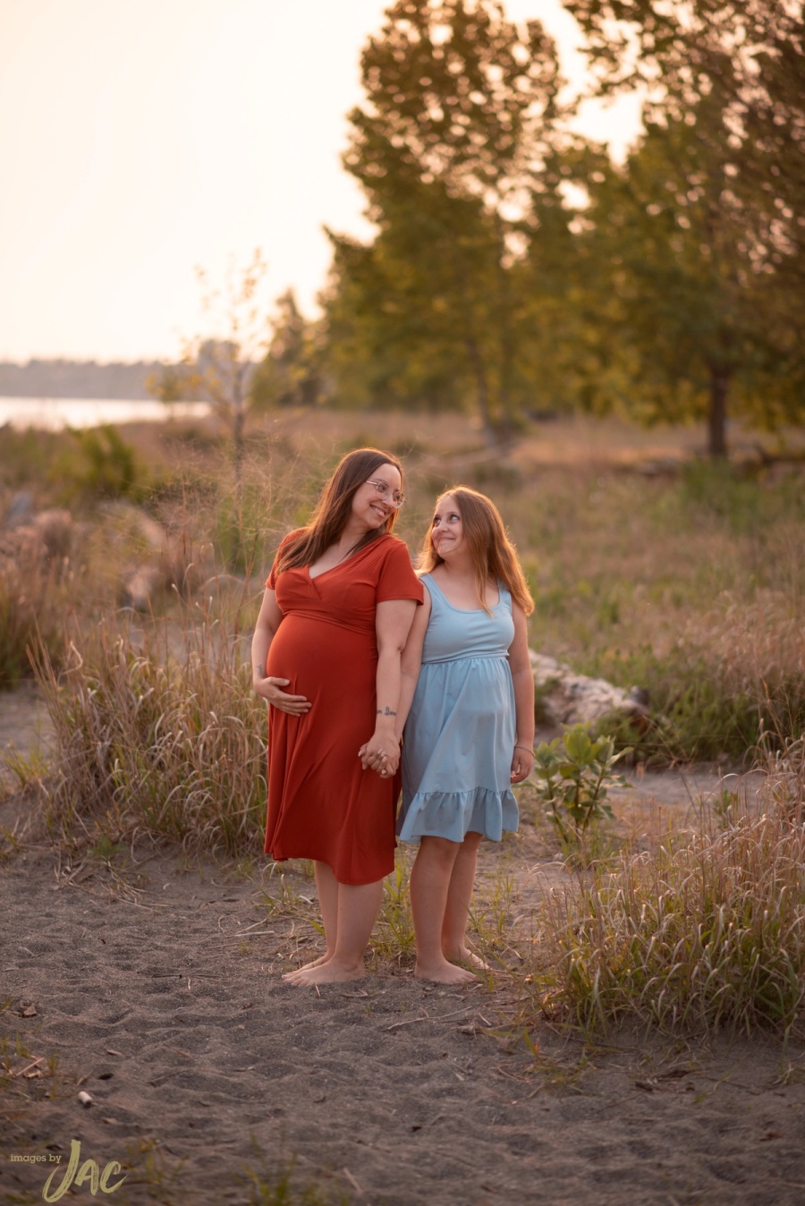A pregnant woman in red and a girl in blue hold hands on the shore at golden hour