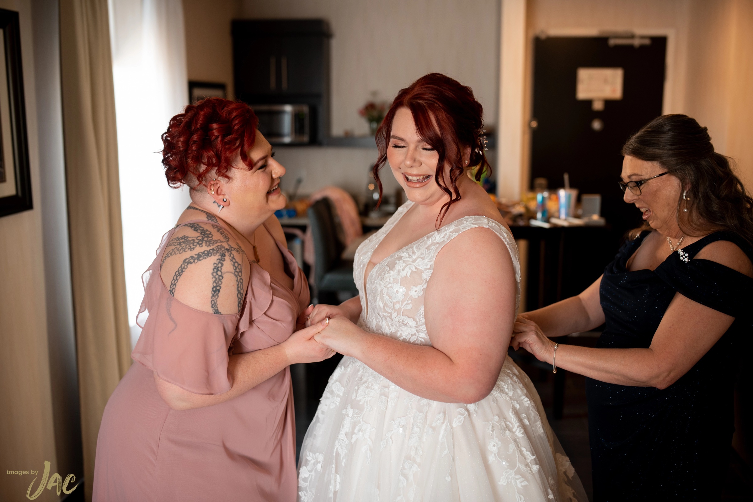 A bride in a lace gown laughs as two women help her into her dress