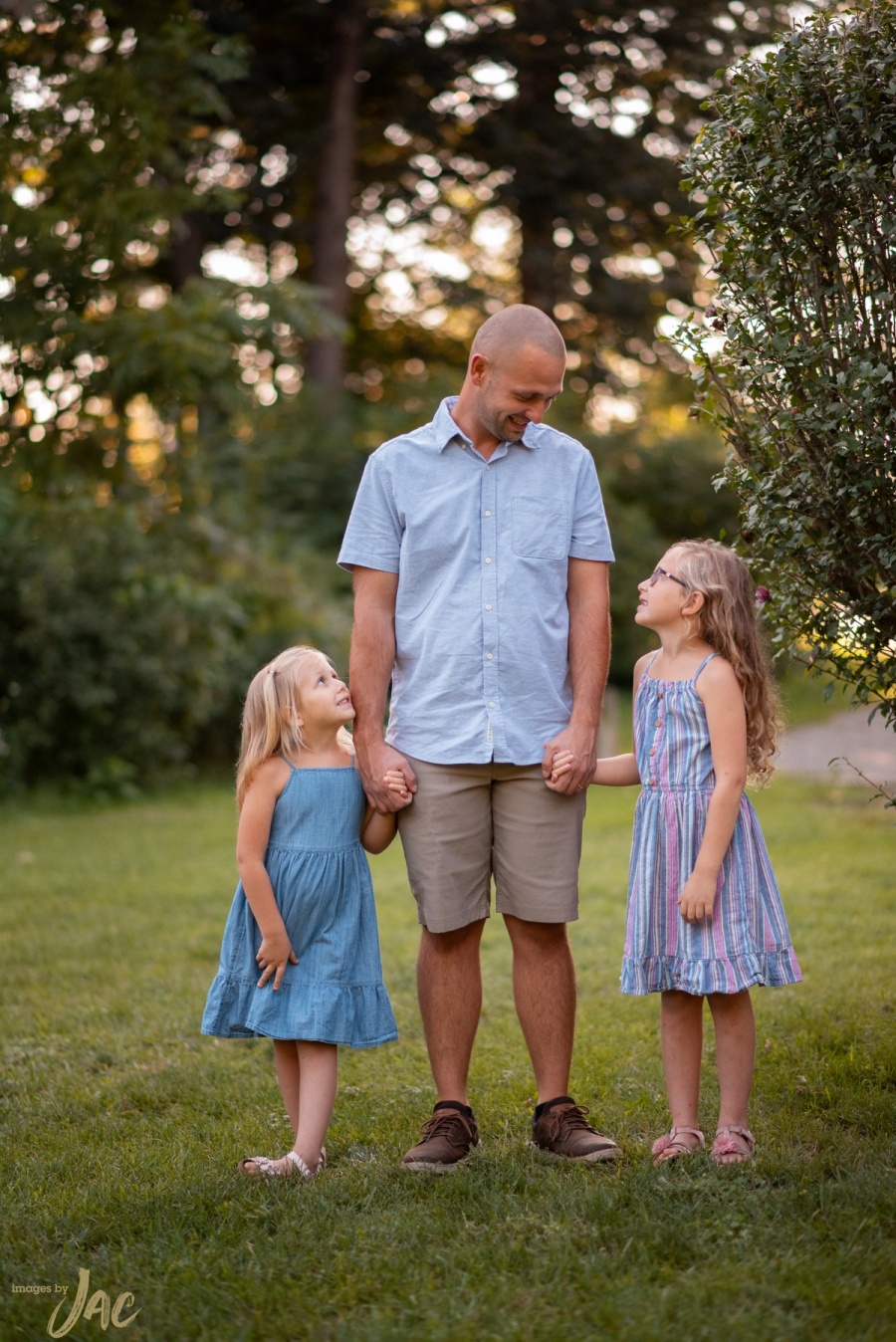 A father walks hand in hand with his two young daughters across a lawn at golden hour