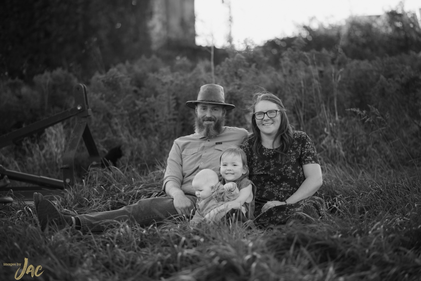 A family sit in tall grass, the father in a hat holding the baby