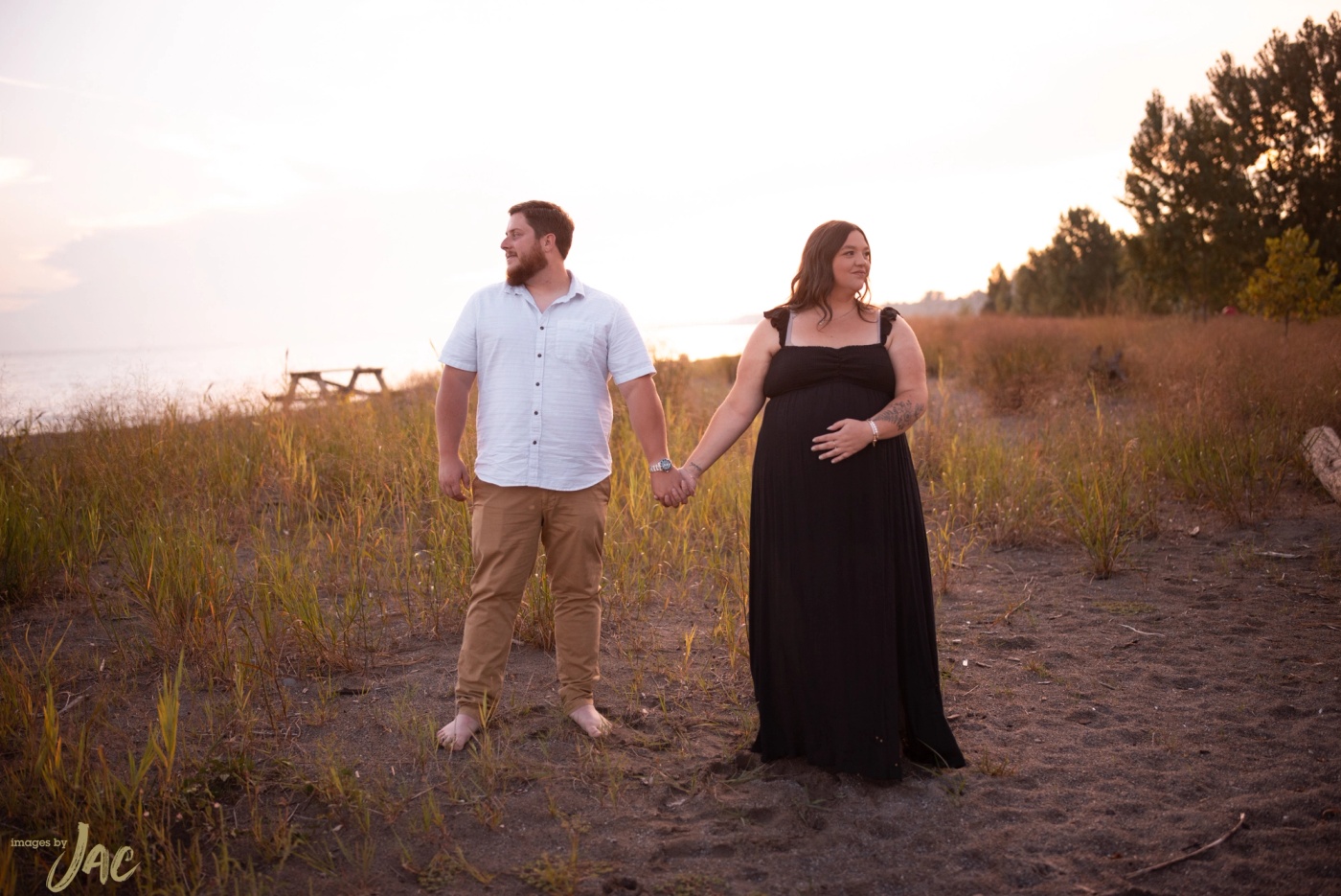 An expectant couple hold hands on a grassy dune at sunset