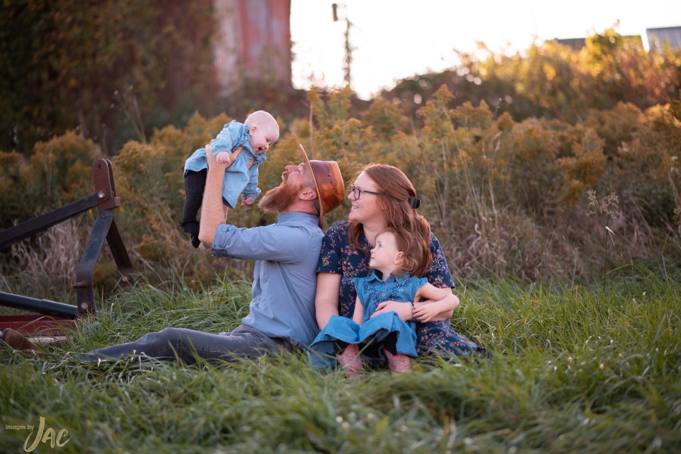 A father lifts a baby overhead as the family sits together in tall grass at sunset