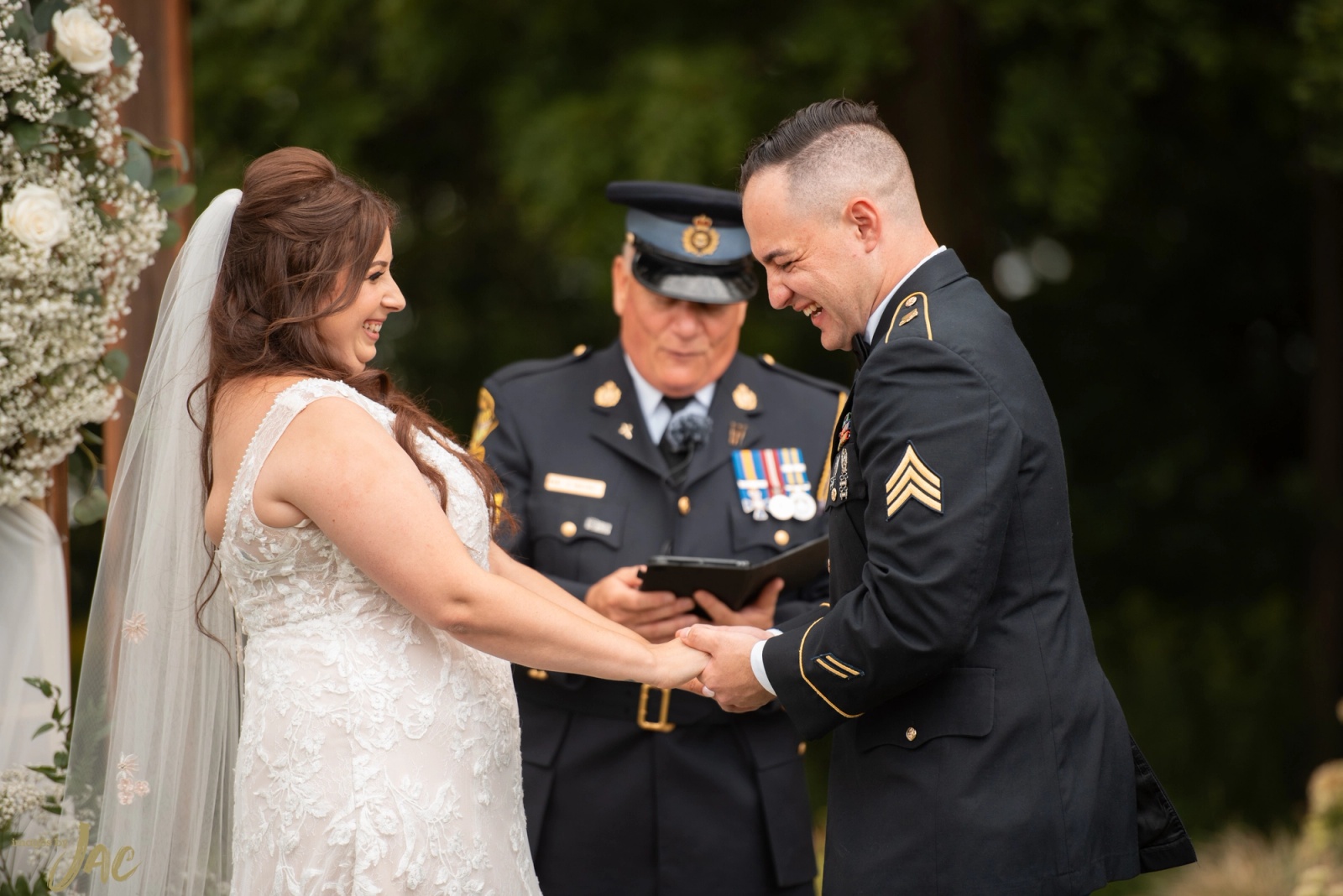 A bride and her groom in military dress uniform laughing as they hold hands during their outdoor ceremony